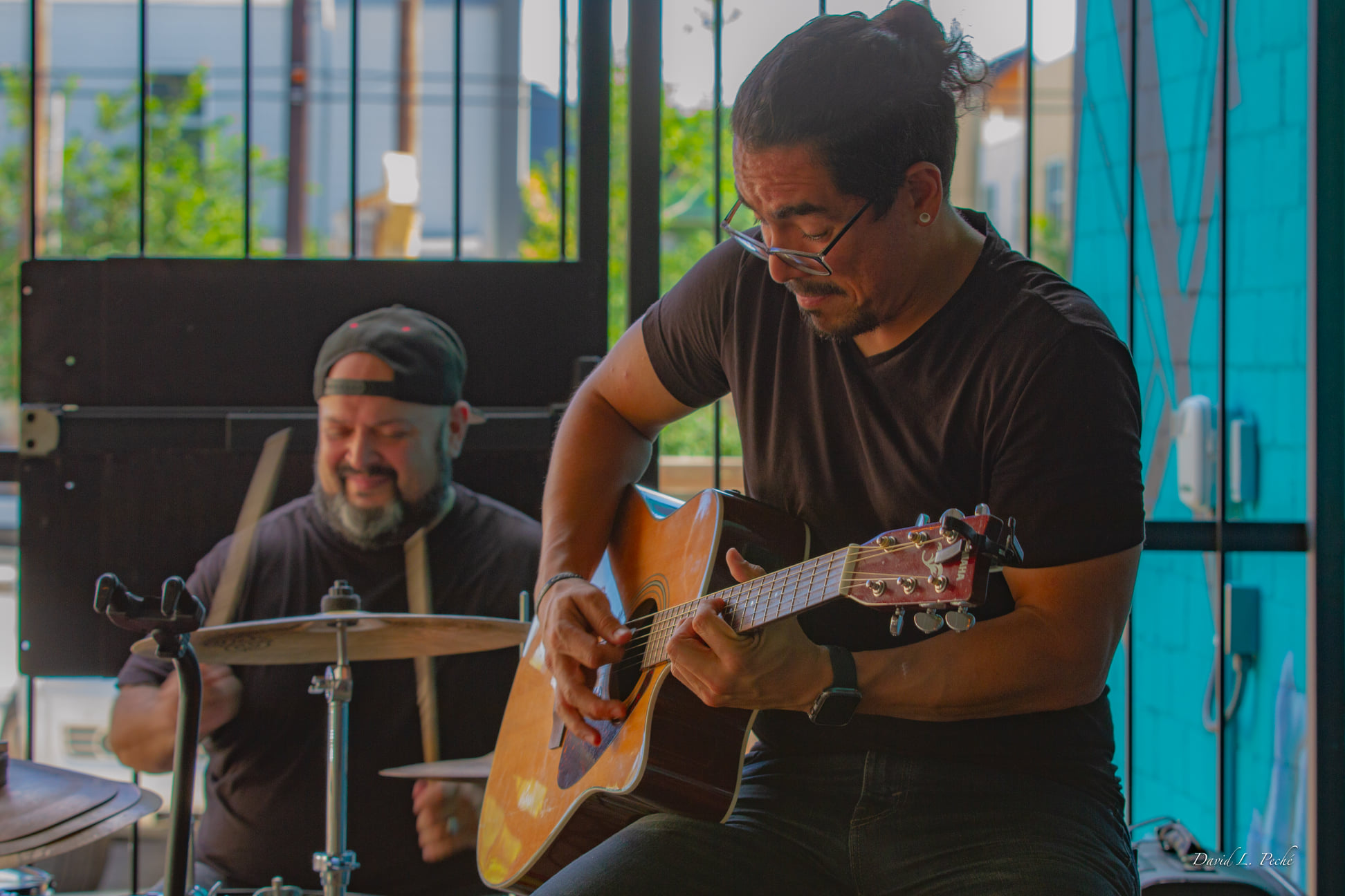 Two musicians perform indoors, one playing guitar and the other on drums, with a window and blue wall in background.