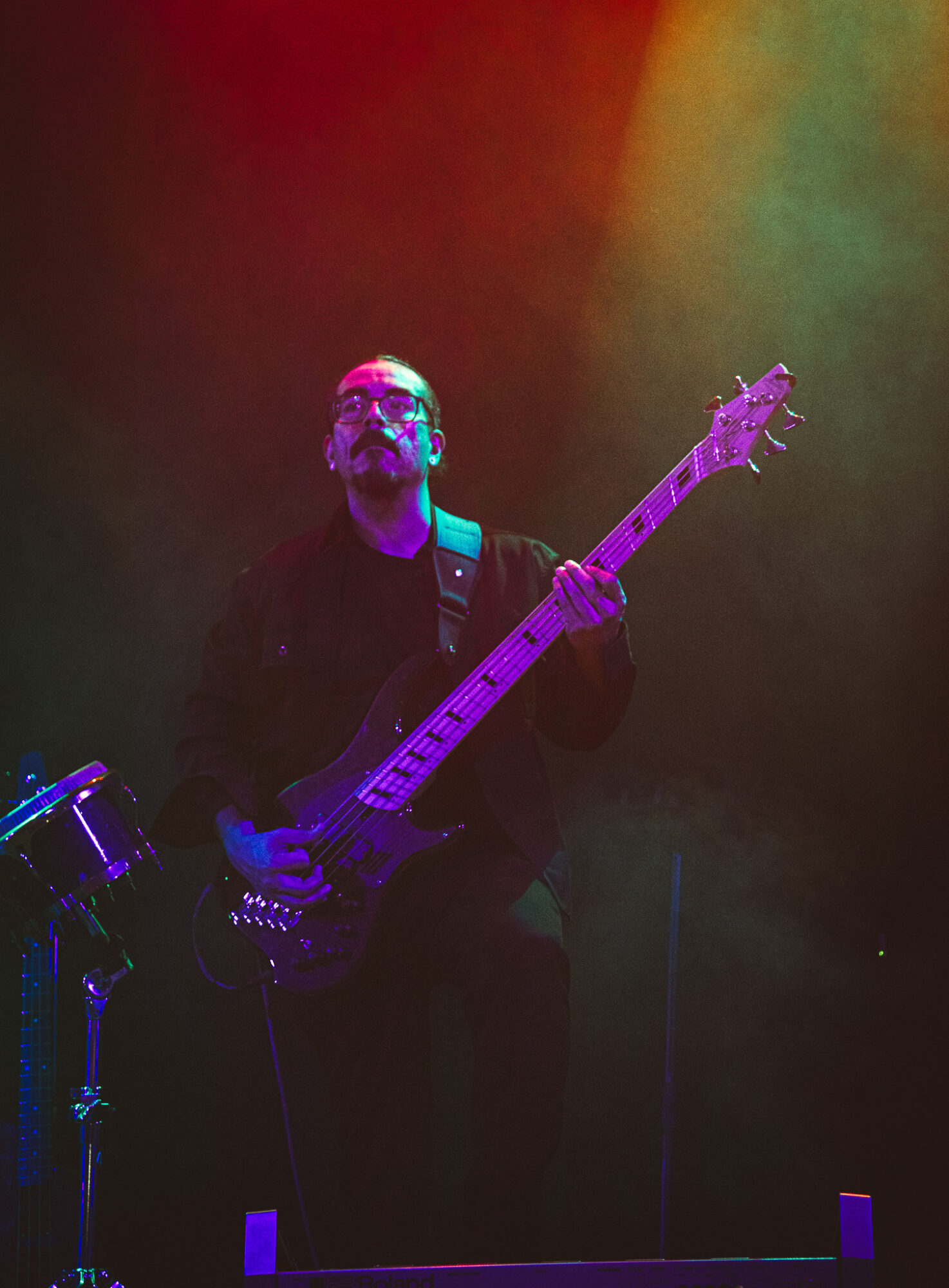 Musician playing bass guitar on stage with colorful lighting, wearing glasses and dark clothing.