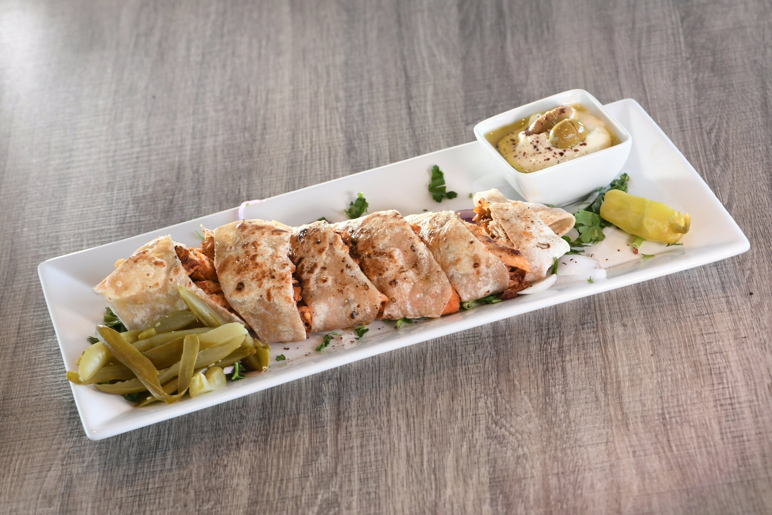 Slices of cooked chicken with pickles and a small bowl of sauce on a rectangular white plate, on a wooden surface.