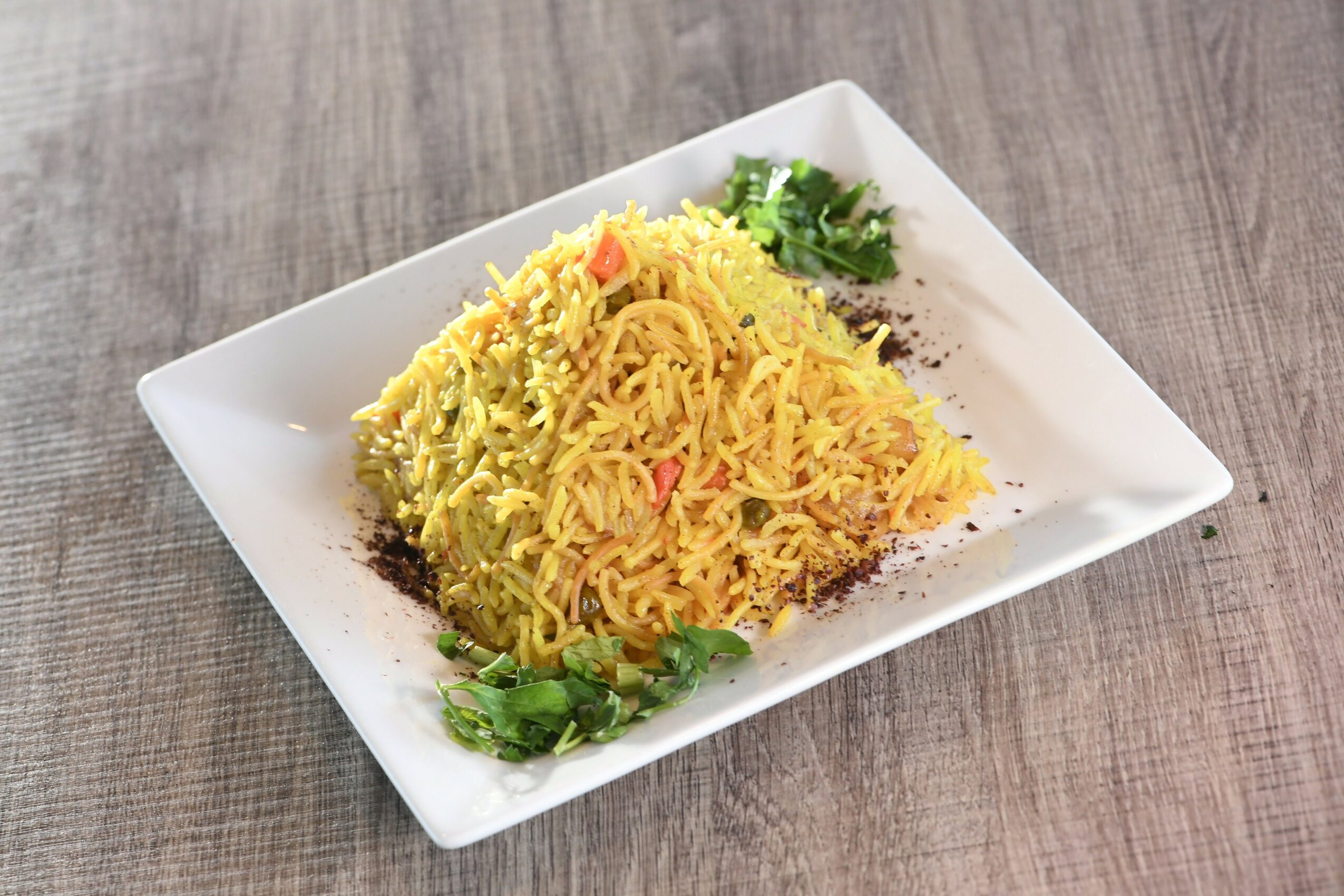 Plate of yellow rice with vegetables garnished with herbs on a white square dish, on a wooden surface.