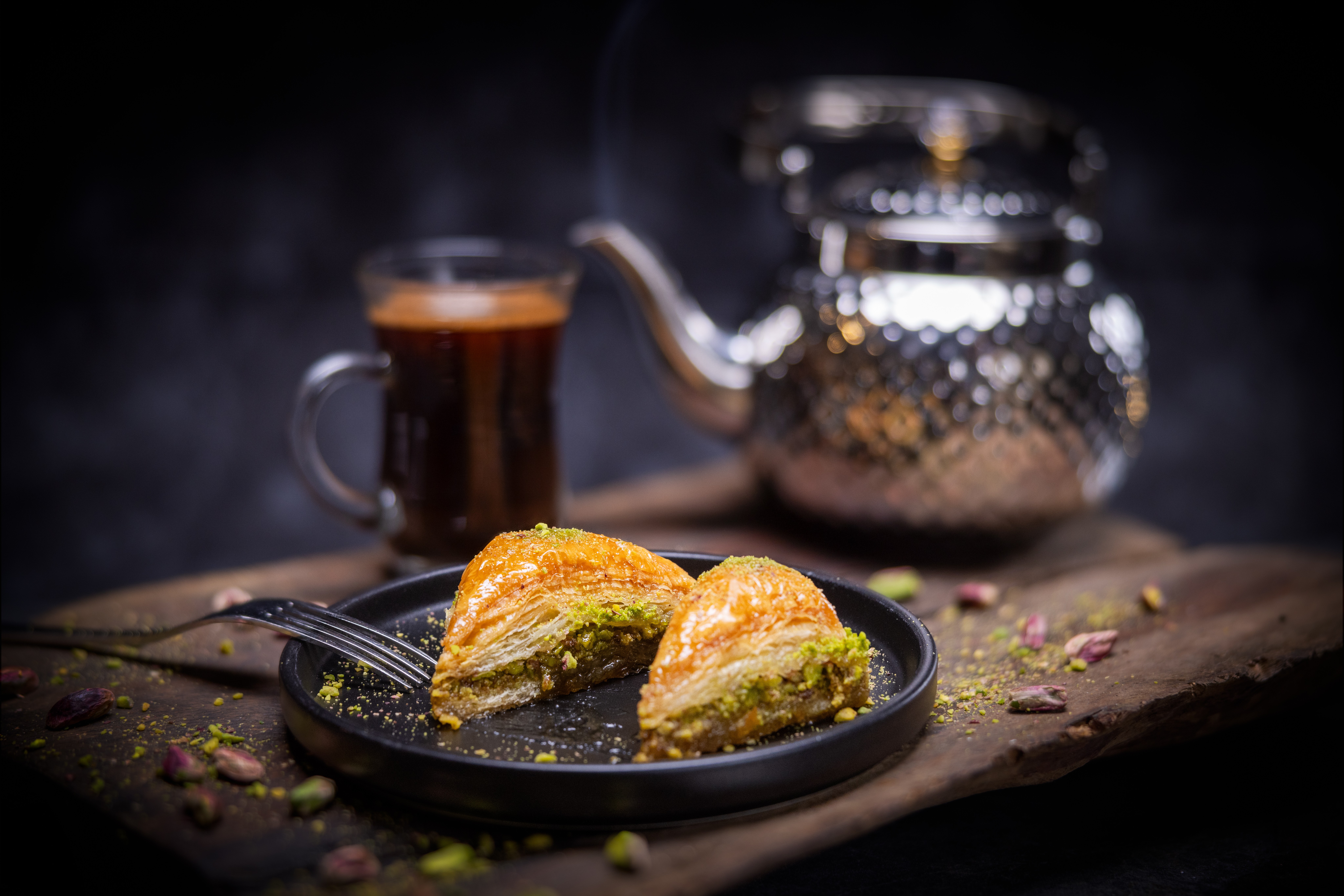 Two slices of cake on a black plate with a teapot and a cup of tea in the background, dark setting.