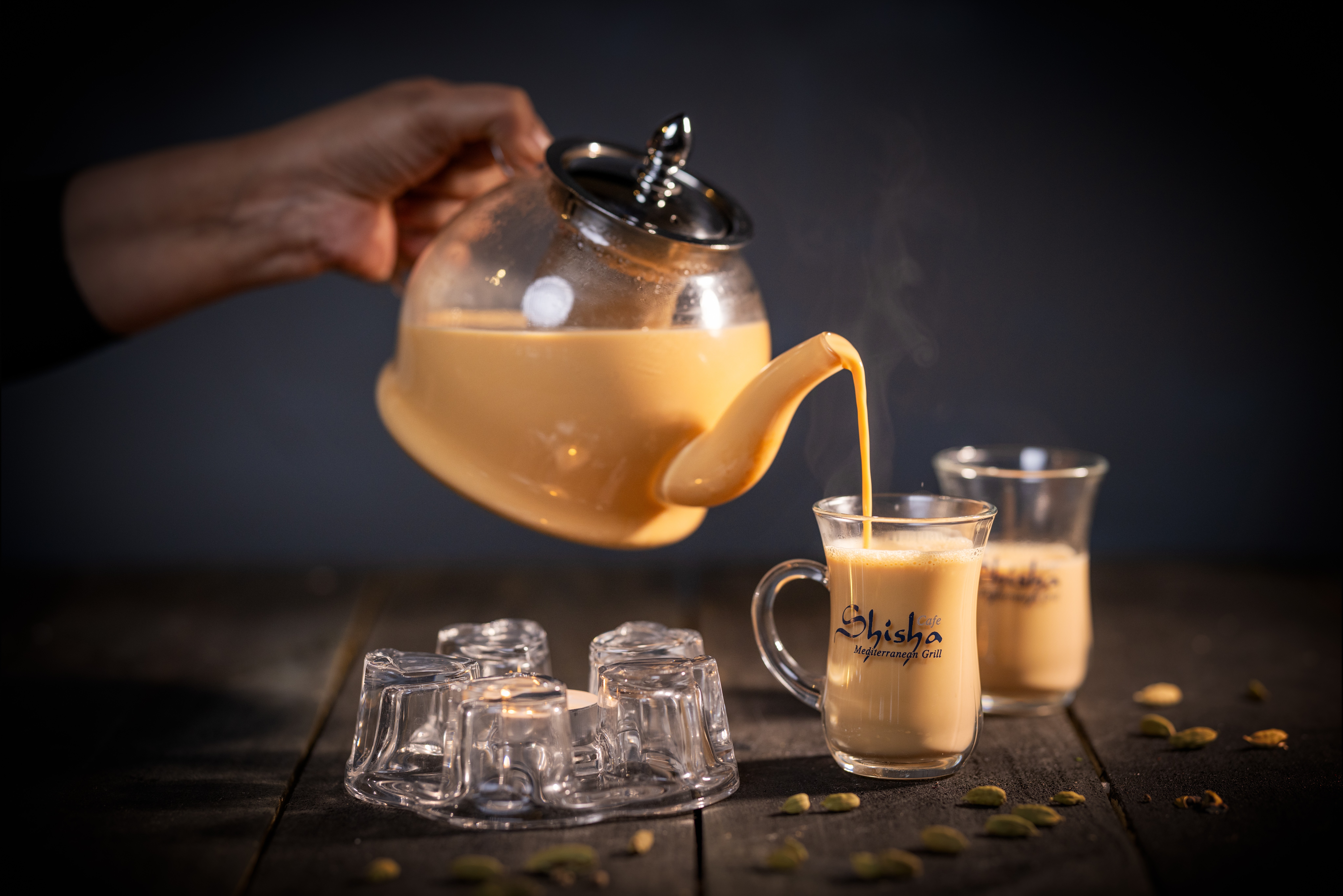 Hand pouring tea from a glass teapot into two cups on a dark surface with ice cubes and scattered seeds, steam rising.