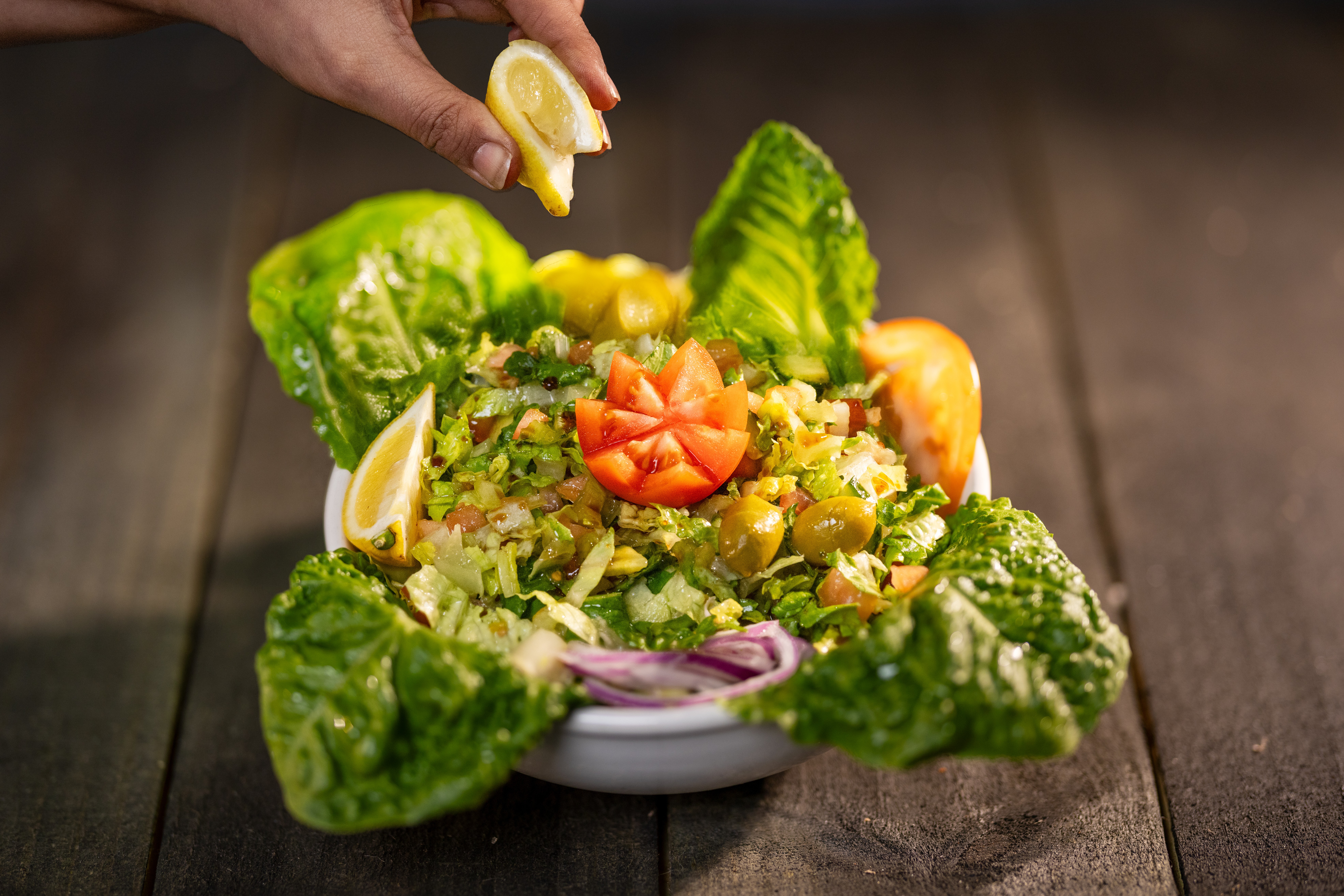 Hand squeezing lemon over a bowl of fresh salad with lettuce, tomatoes, and vegetables on a wooden surface.