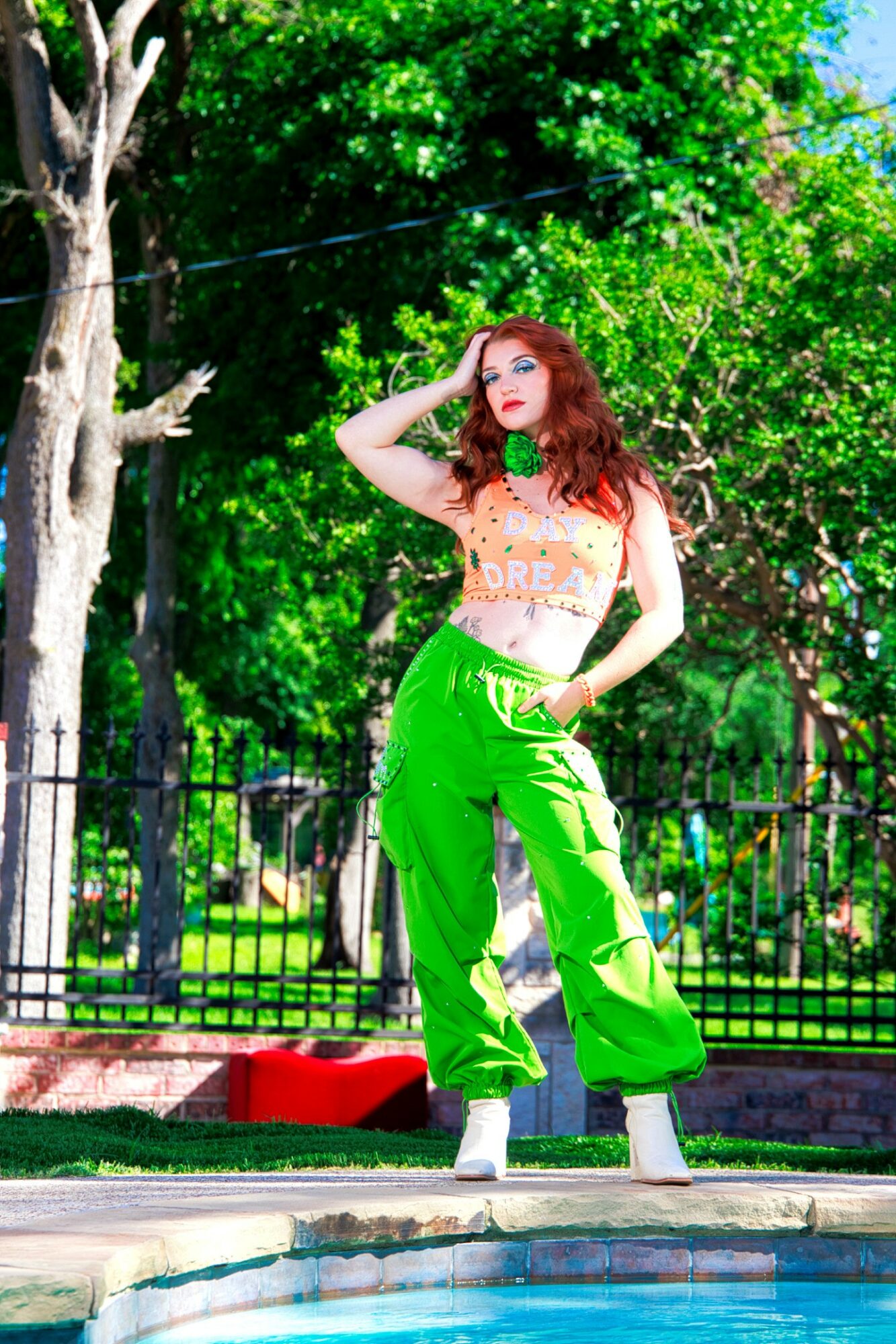 Woman with red hair in green pants and a patterned top stands outdoors near a pool, with trees and a fence in background.