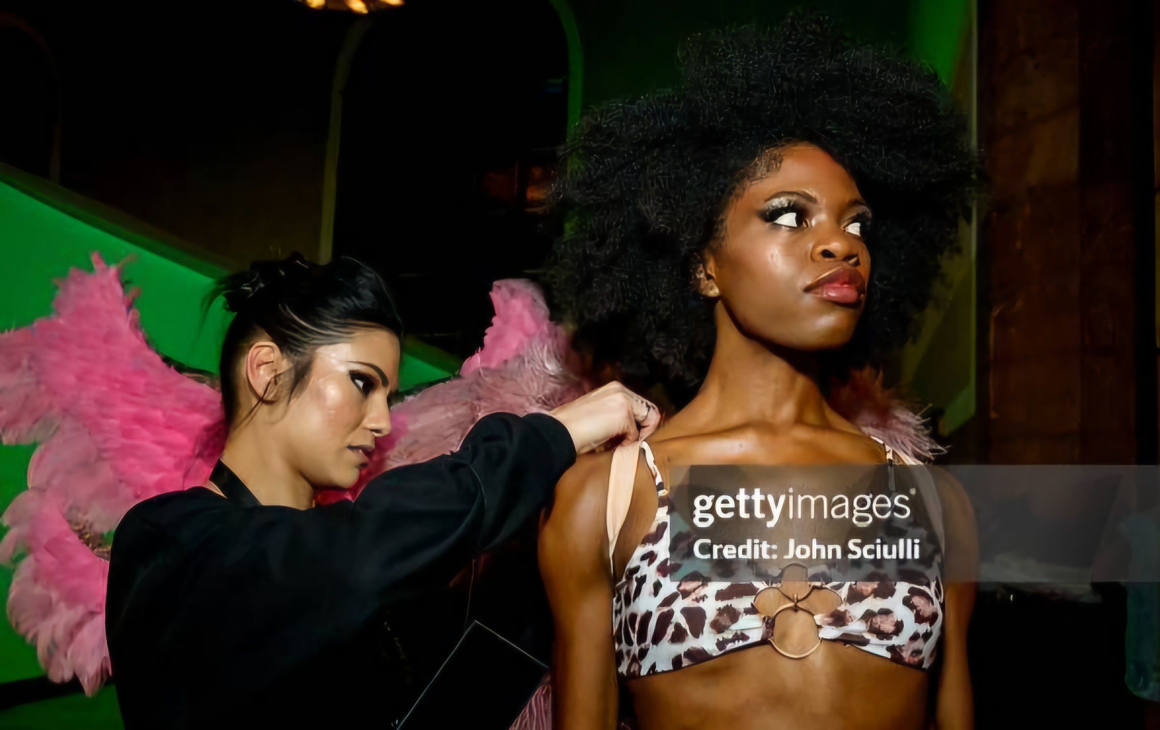Two women, one with large curly hair and leopard print top, the other with dark hair and pink feathered wings, adjusting the first woman's strap.