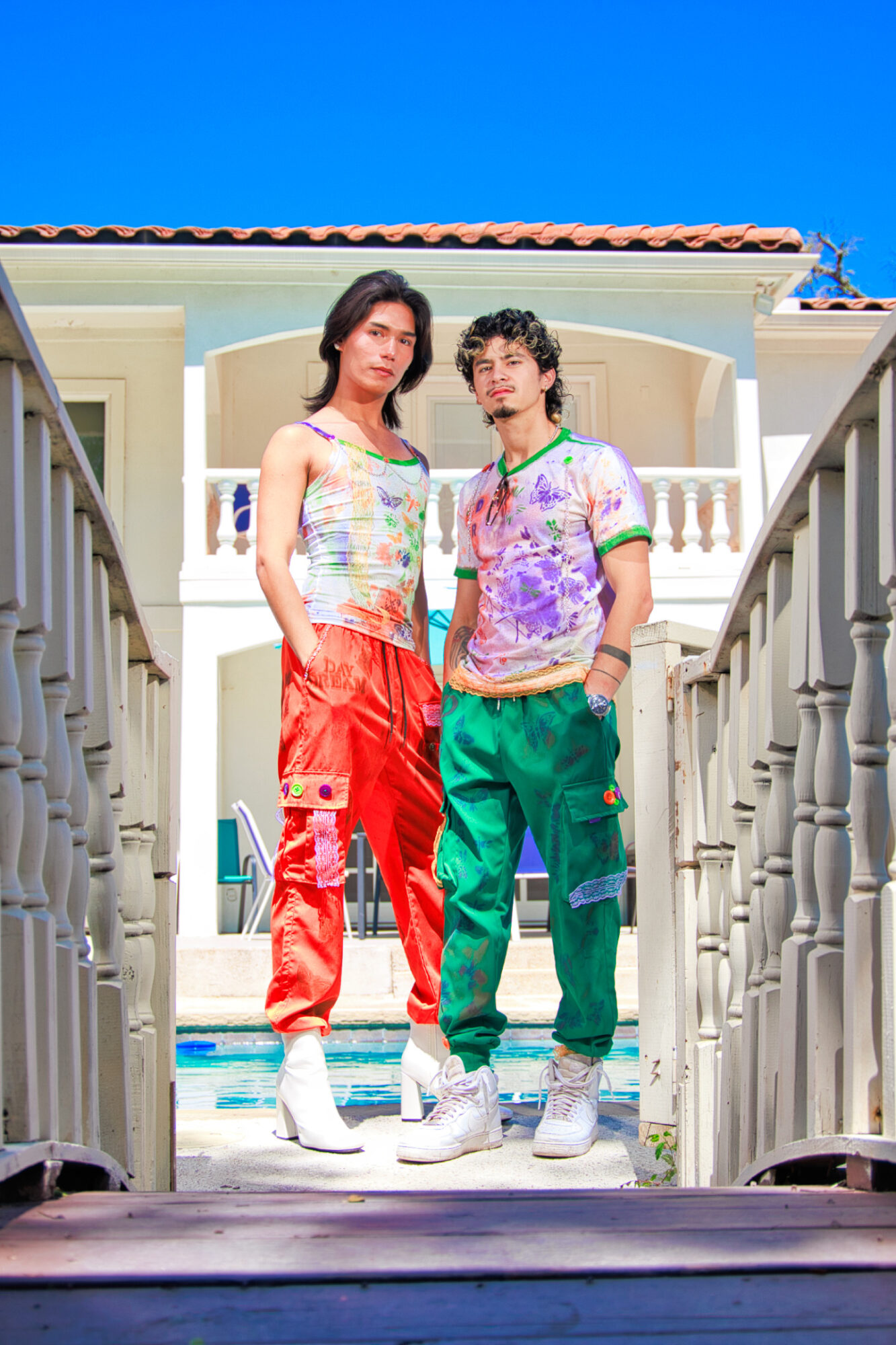 Two young people stand on a staircase outdoors, with a building and blue sky in the background.