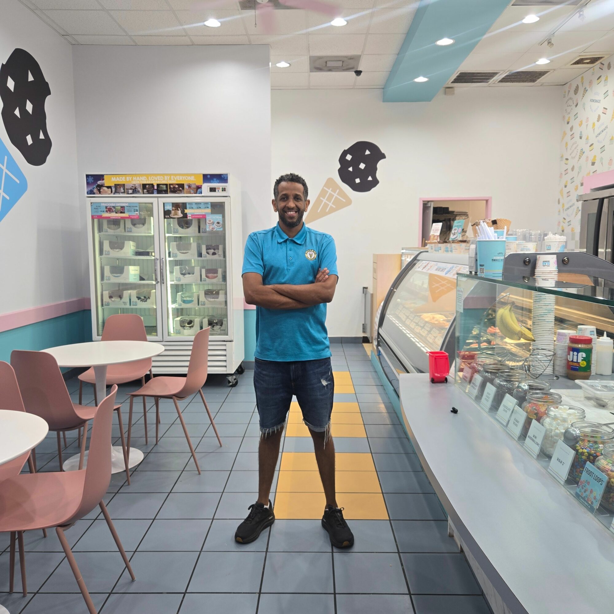 A person in a blue shirt stands with arms crossed in a store, with a display case and pink chairs nearby.