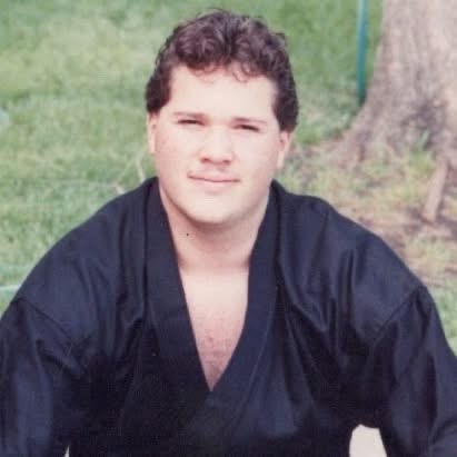 Young man with curly hair in a black martial arts uniform outdoors, near a tree and grass.