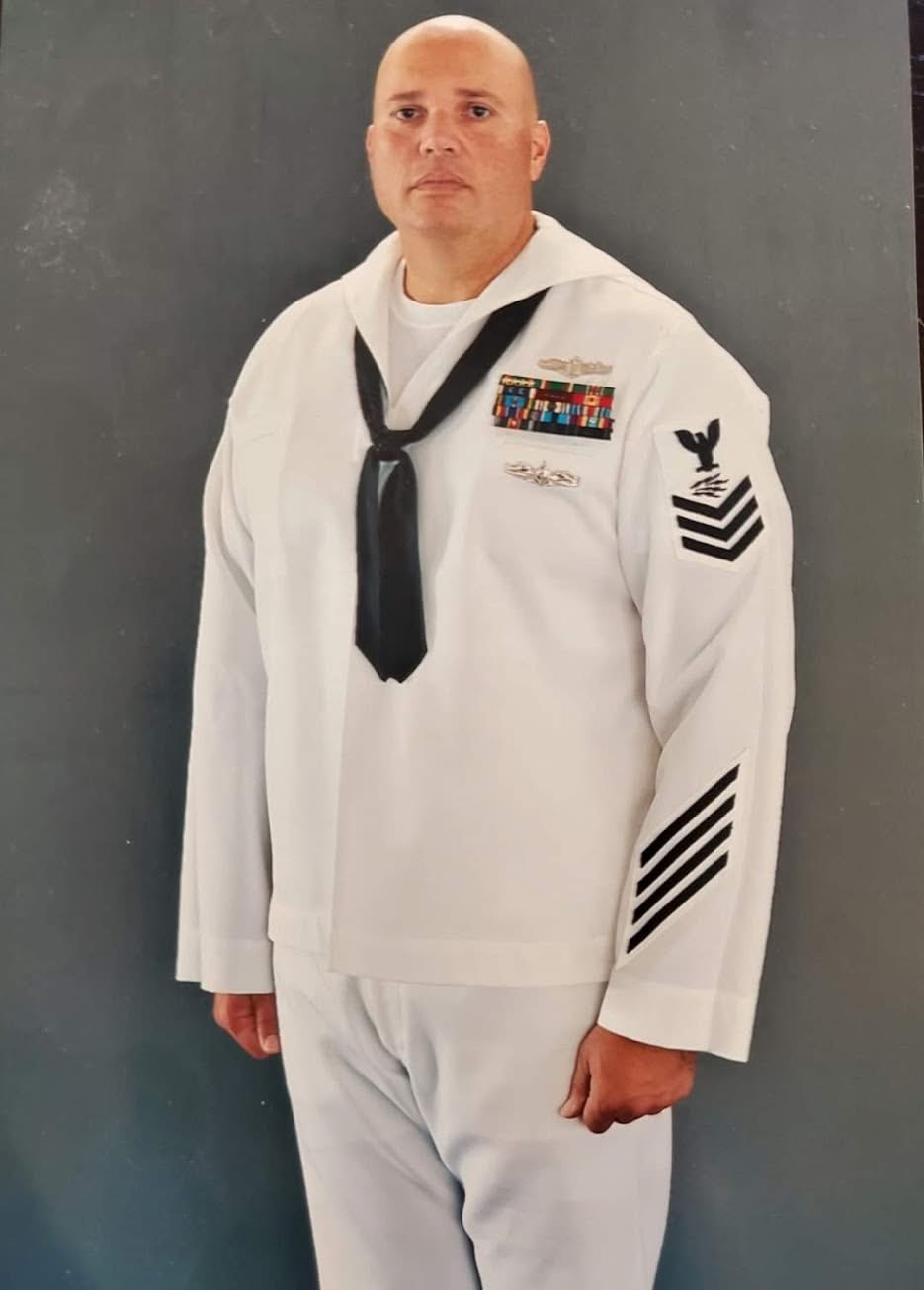 Man in a white naval uniform with medals, black neckerchief, and rank insignia, standing against a dark background.