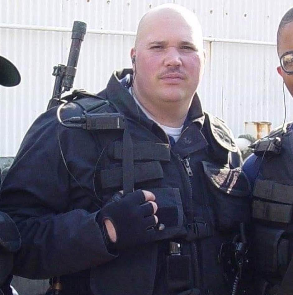 Bald man in police uniform with tactical gear, standing outdoors with a light-colored fence in background.