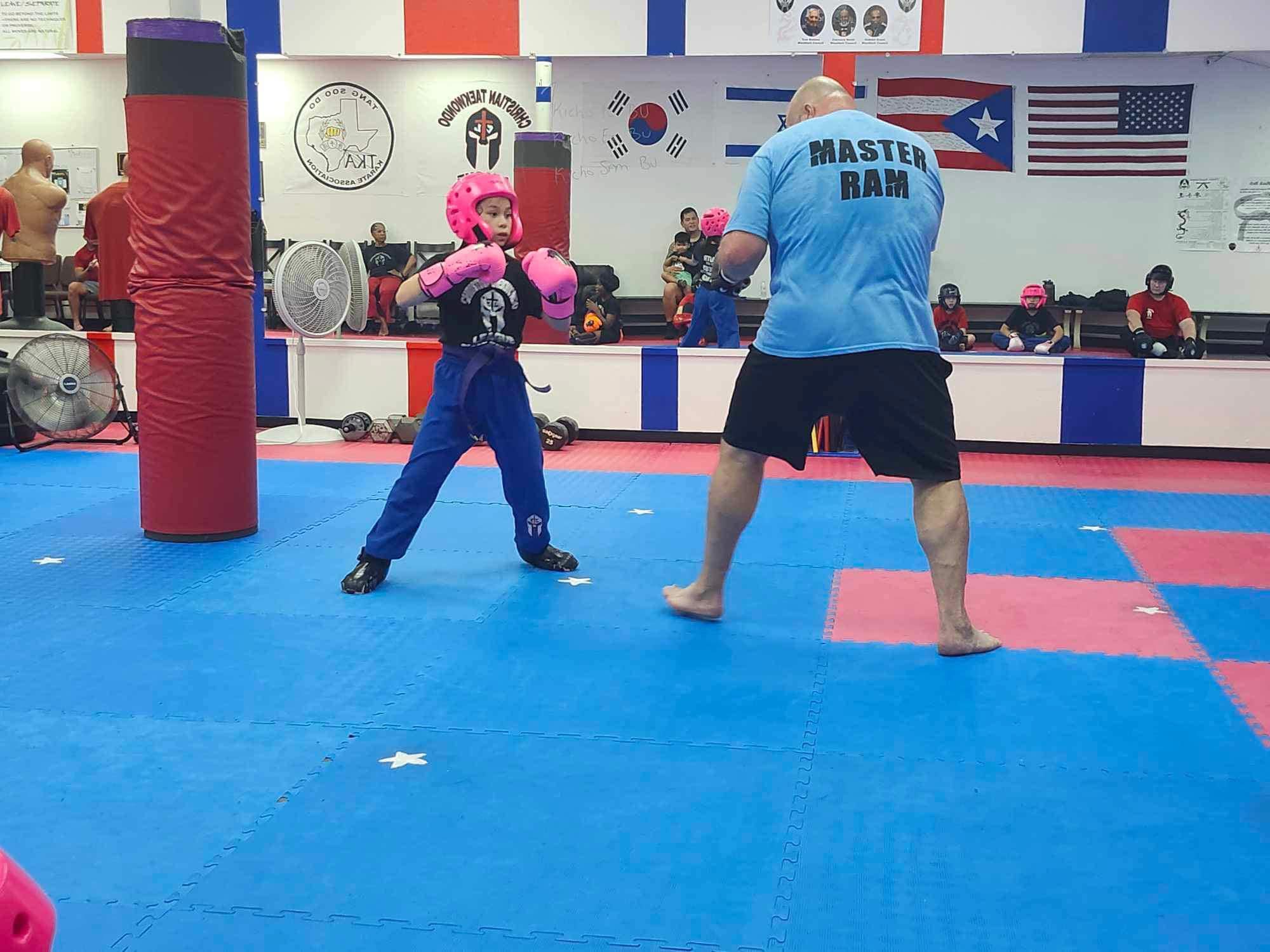 Two people practicing martial arts in a gym with flags on the wall, one in pink gloves and blue uniform, the other in a blue shirt and black shorts.