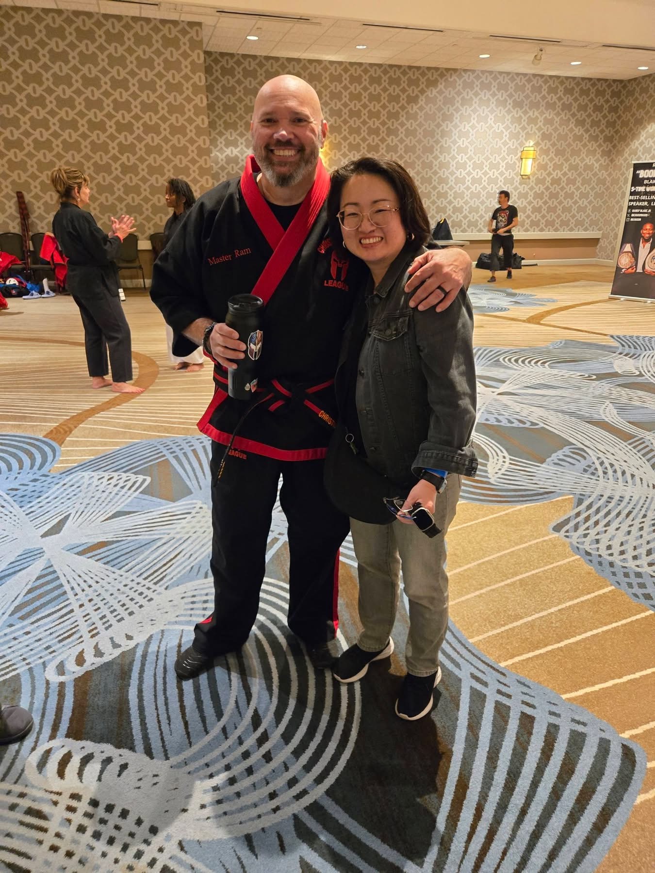 Two people smiling and posing together indoors, one with arm around the other, with a patterned carpet and other people in background.