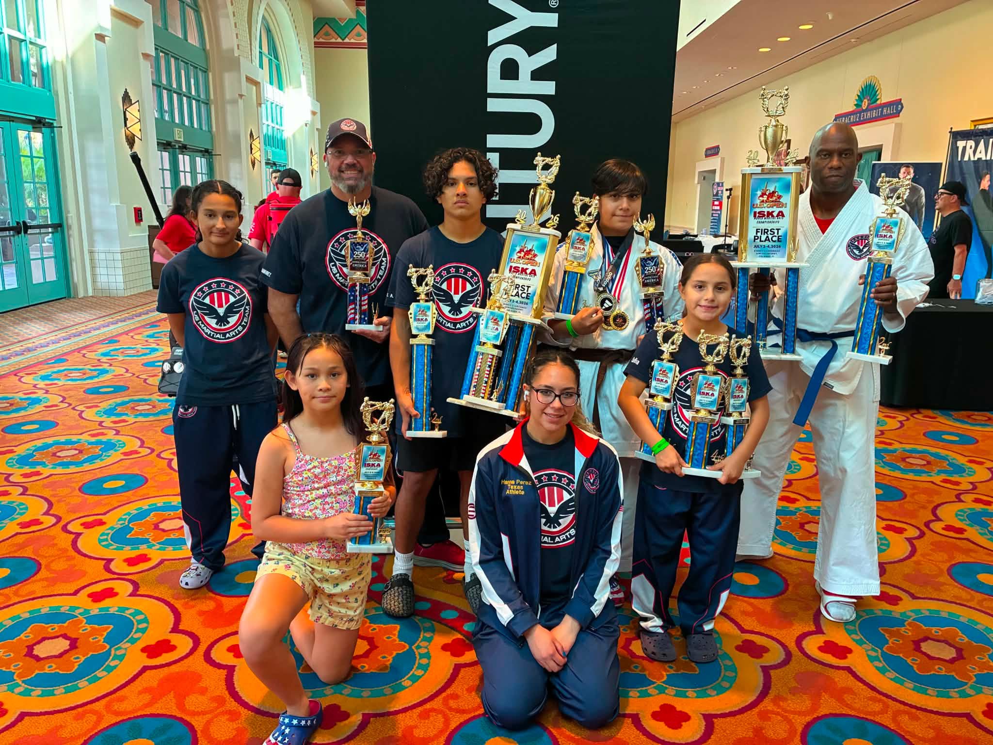 Group of children and adults holding trophies in a colorful indoor space with banners and displays.
