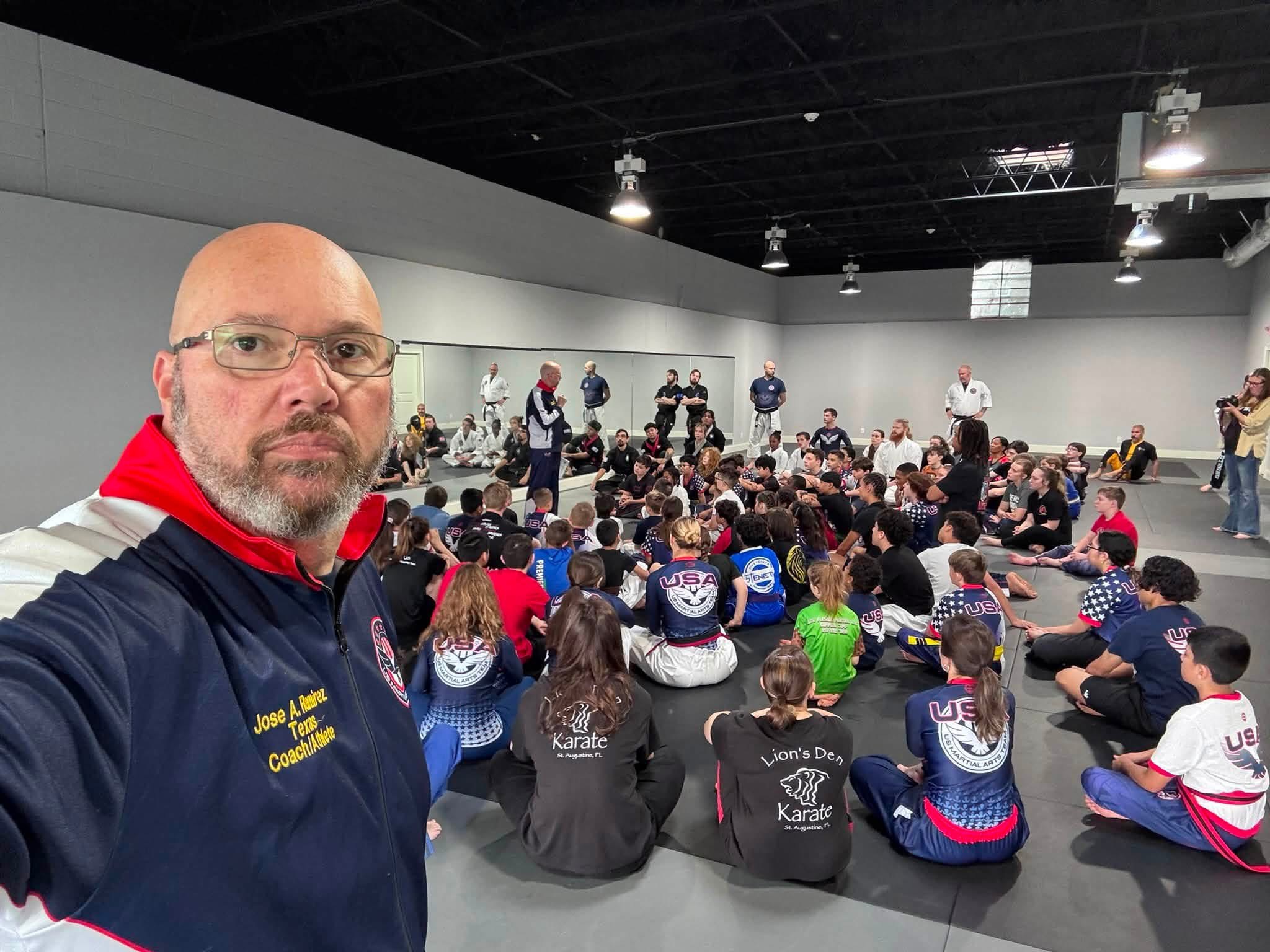 Man with glasses and beard wearing a dark jacket with red collar, in front of a large group of children sitting on the floor in a spacious room.