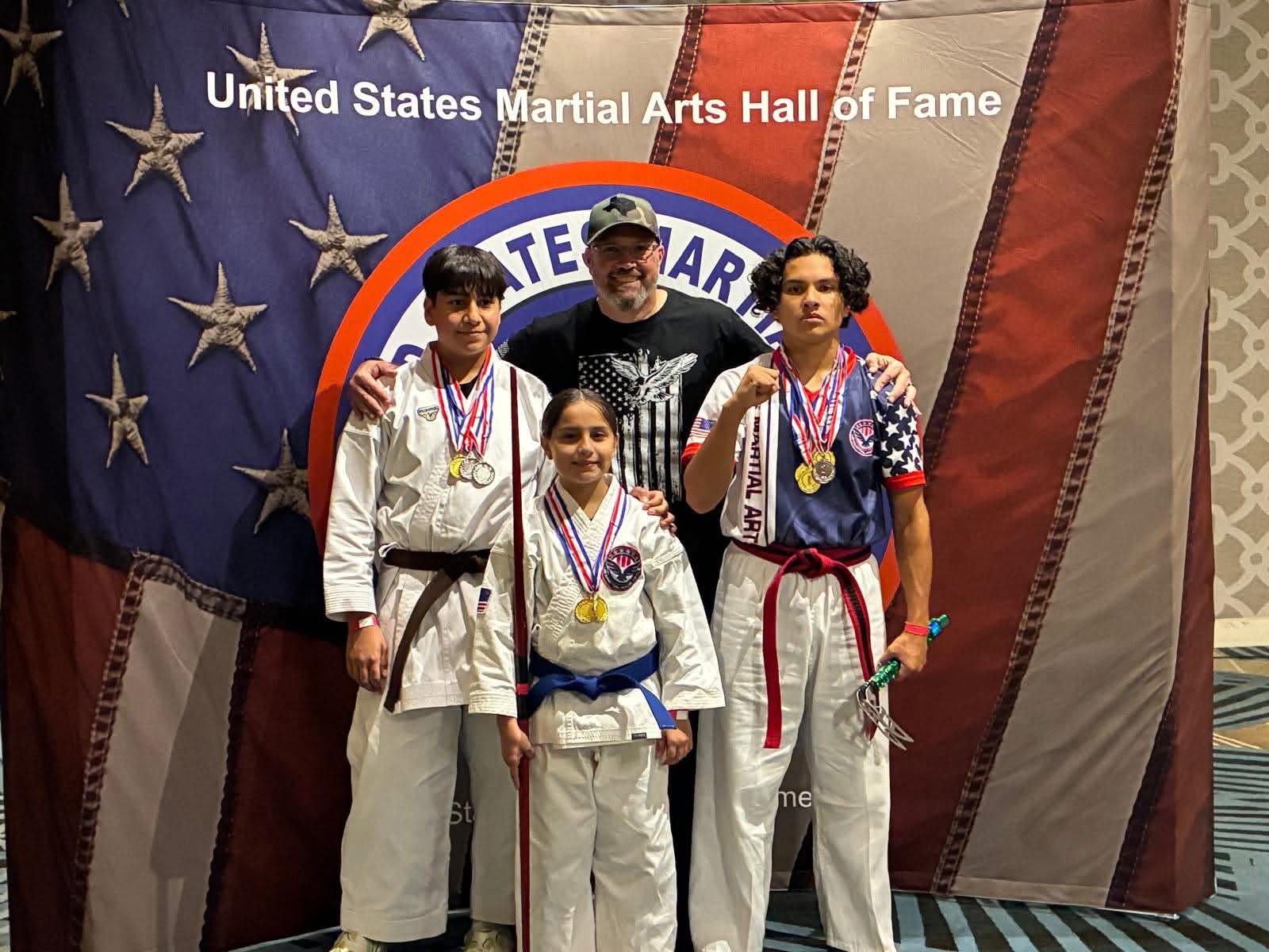 Four martial artists in uniforms with medals, standing in front of a United States flag and martial arts hall of fame backdrop.