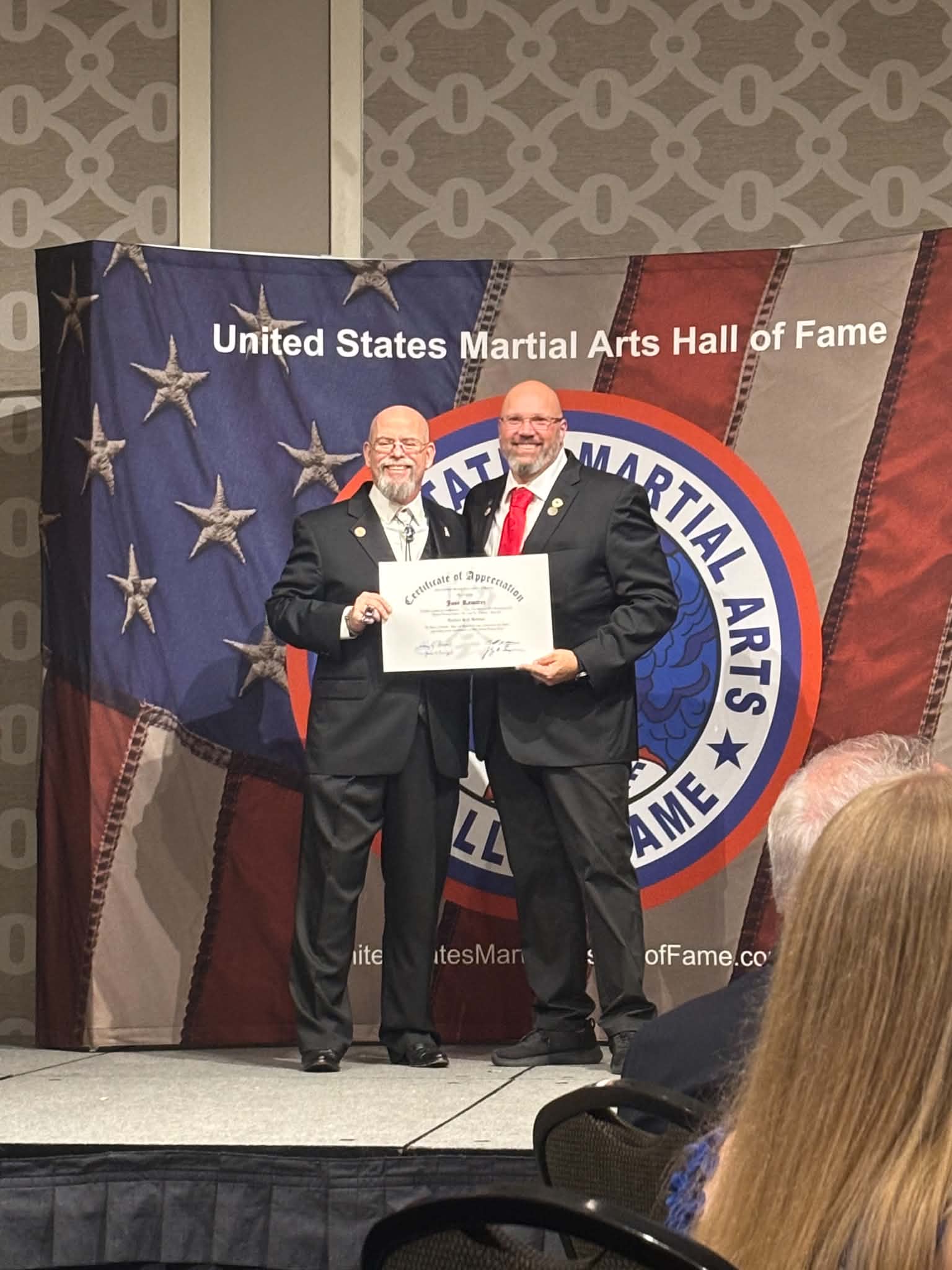 Two men in suits holding a certificate on stage with a United States Martial Arts Hall of Fame banner behind them.