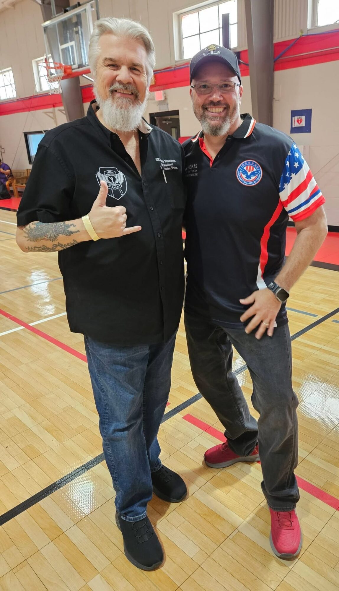 Two men standing in a gymnasium, smiling, with one making a hand gesture, both wearing casual and sports attire.