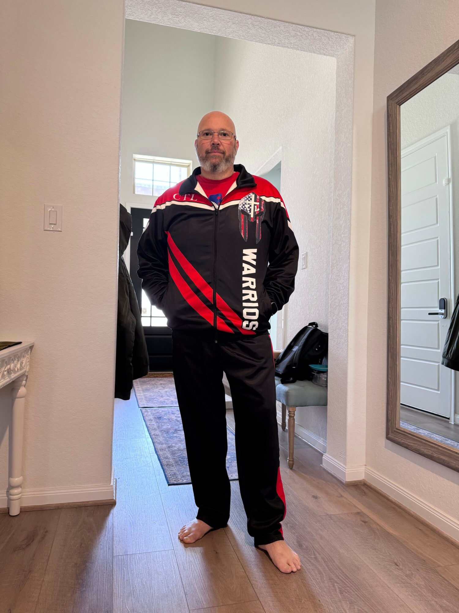 Person standing in a hallway, wearing a black and red sports jacket and black pants, with a door and window in the background.