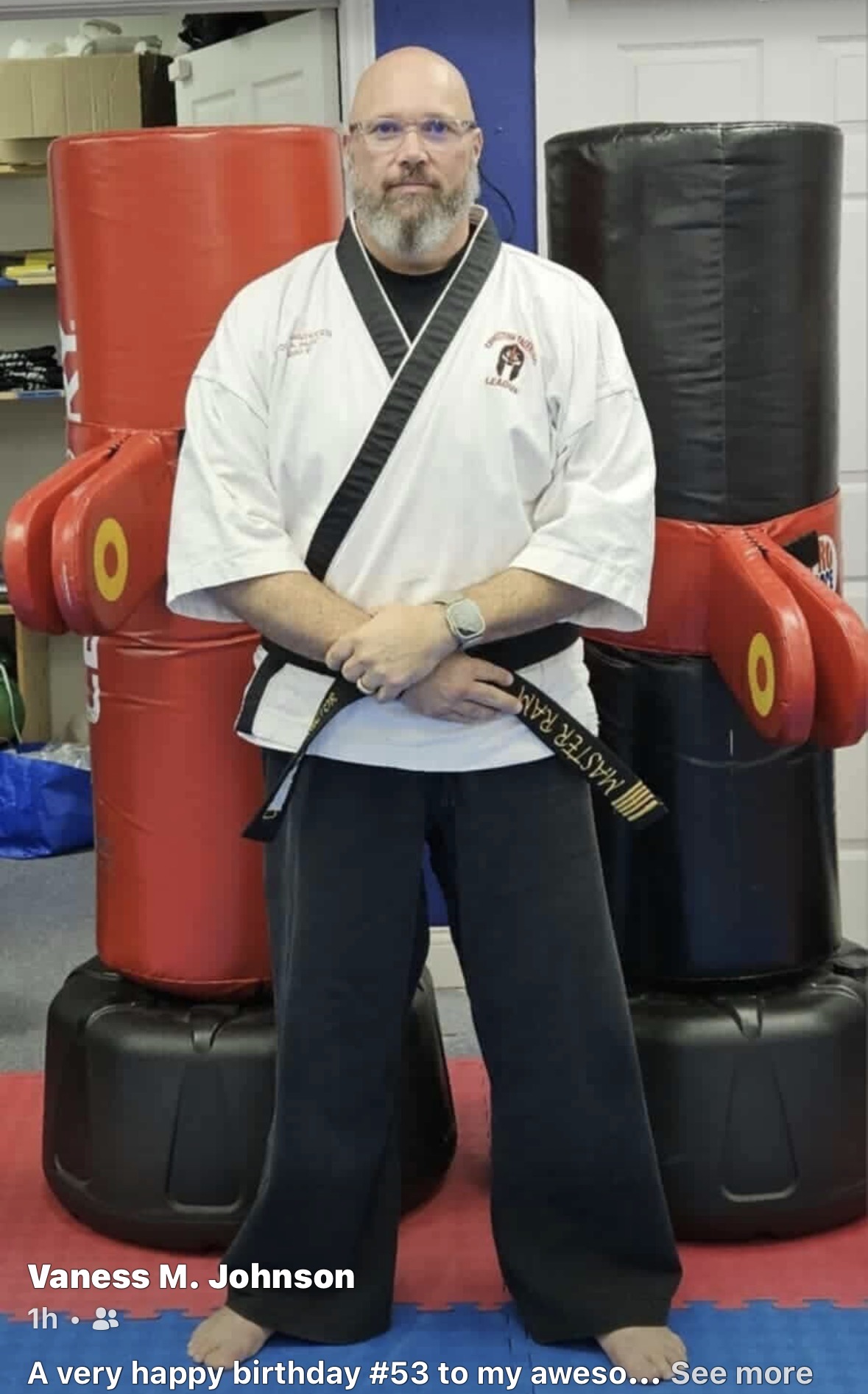 Man in martial arts uniform standing in front of padded training equipment, holding a black belt, in a training room.