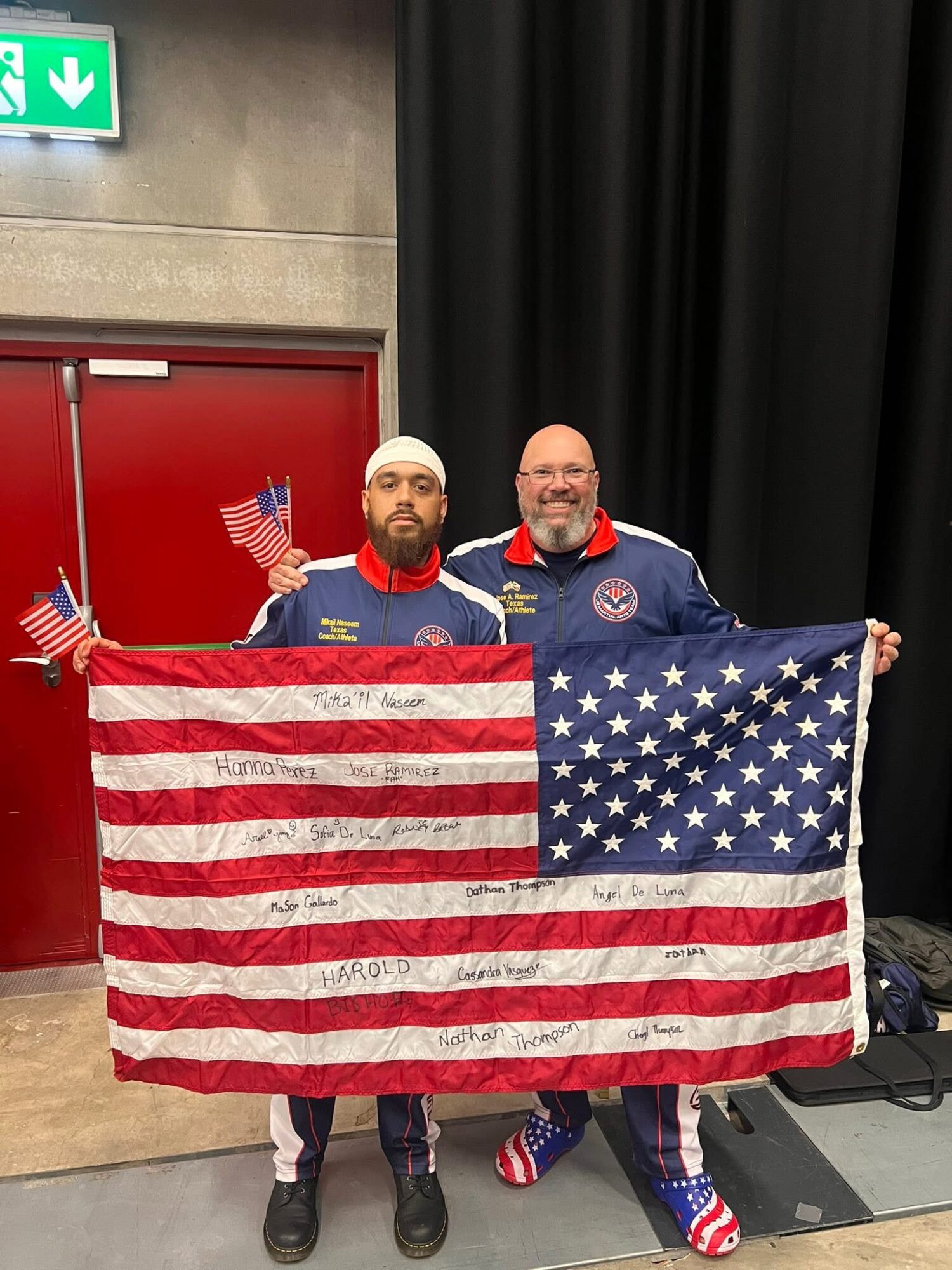 Two men in uniforms holding an American flag with handwritten messages, standing indoors with black curtains behind them.