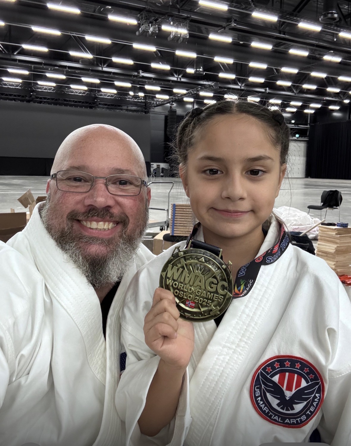 A man and a girl in martial arts uniforms holding a medal, smiling indoors with bright ceiling lights.