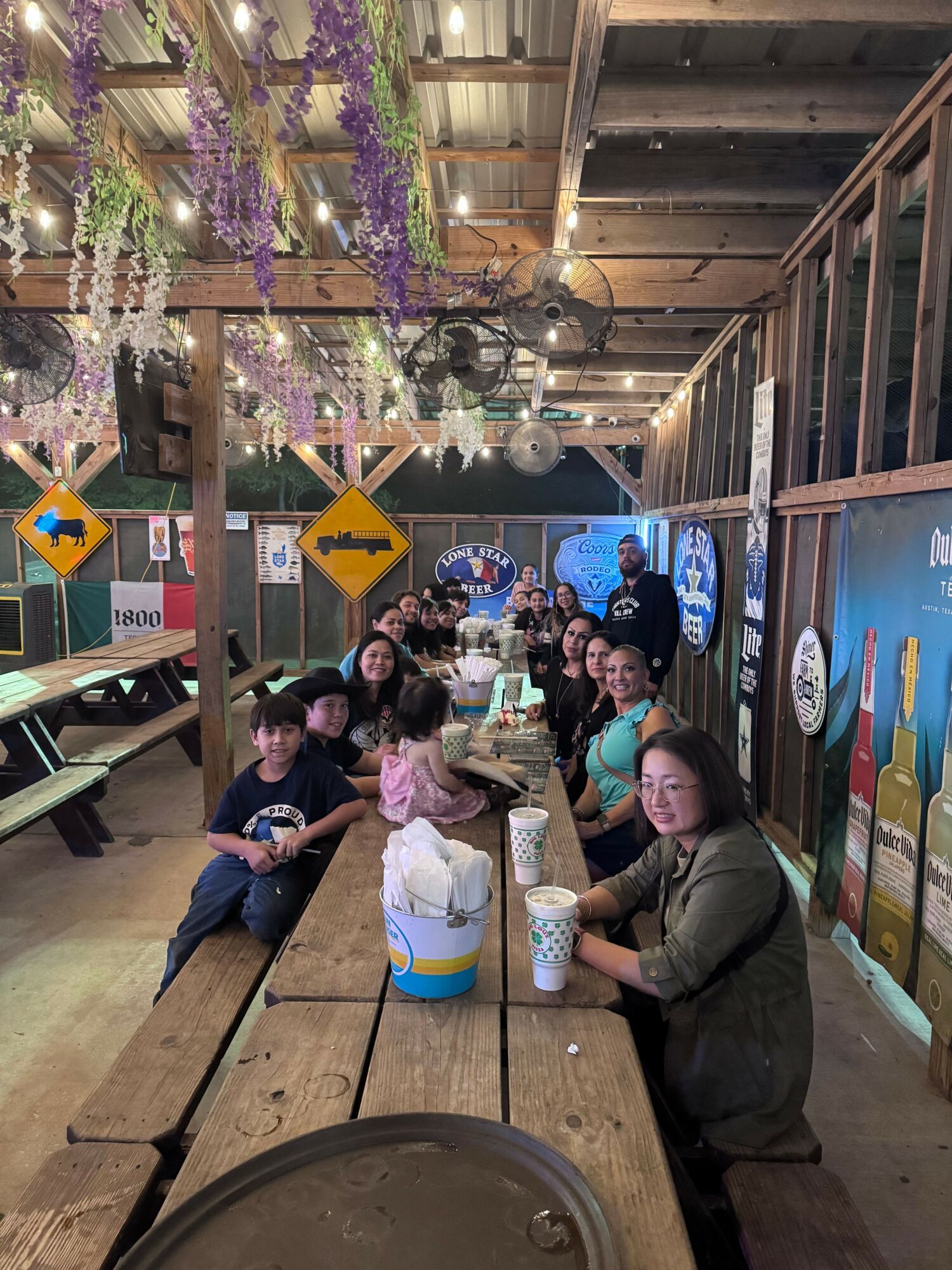 Group of people sitting on a wooden bench at an outdoor area with hanging purple flowers and string lights, some holding drinks.