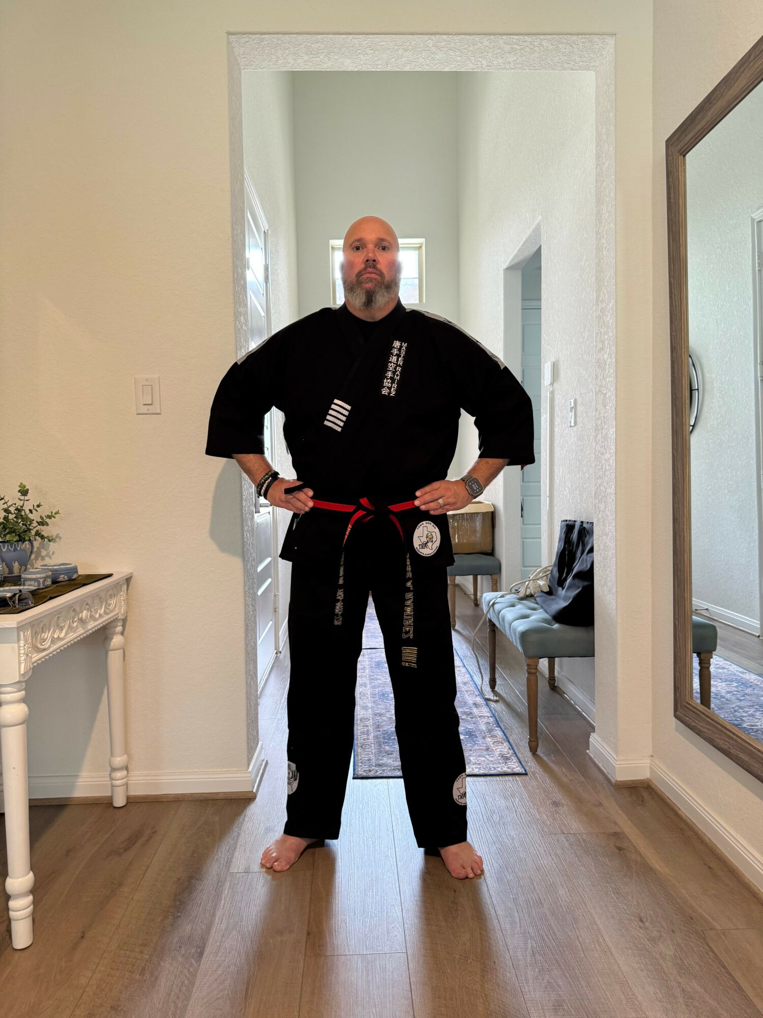 Person in black martial arts uniform with red belt standing in a room, holding a small object, with a doorway and furniture visible.