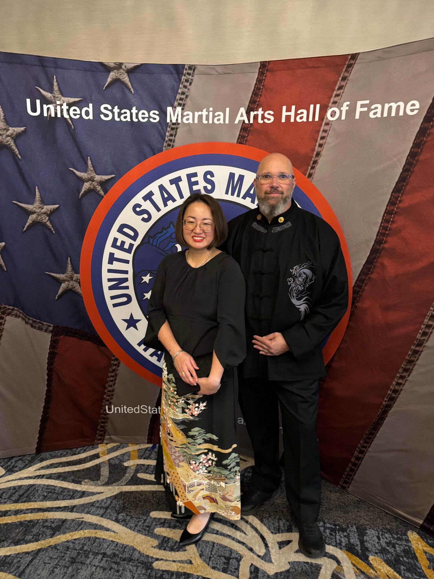 Two women stand in front of a United States Martial Arts Hall of Fame backdrop, one holding a certificate, both smiling.