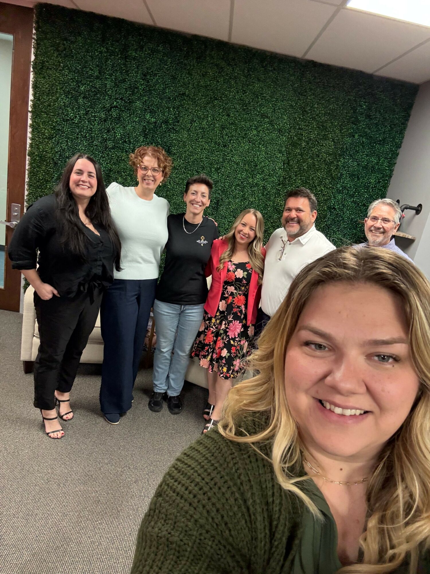 Group of six people standing in front of a green wall, taking a selfie with a woman in the foreground smiling.