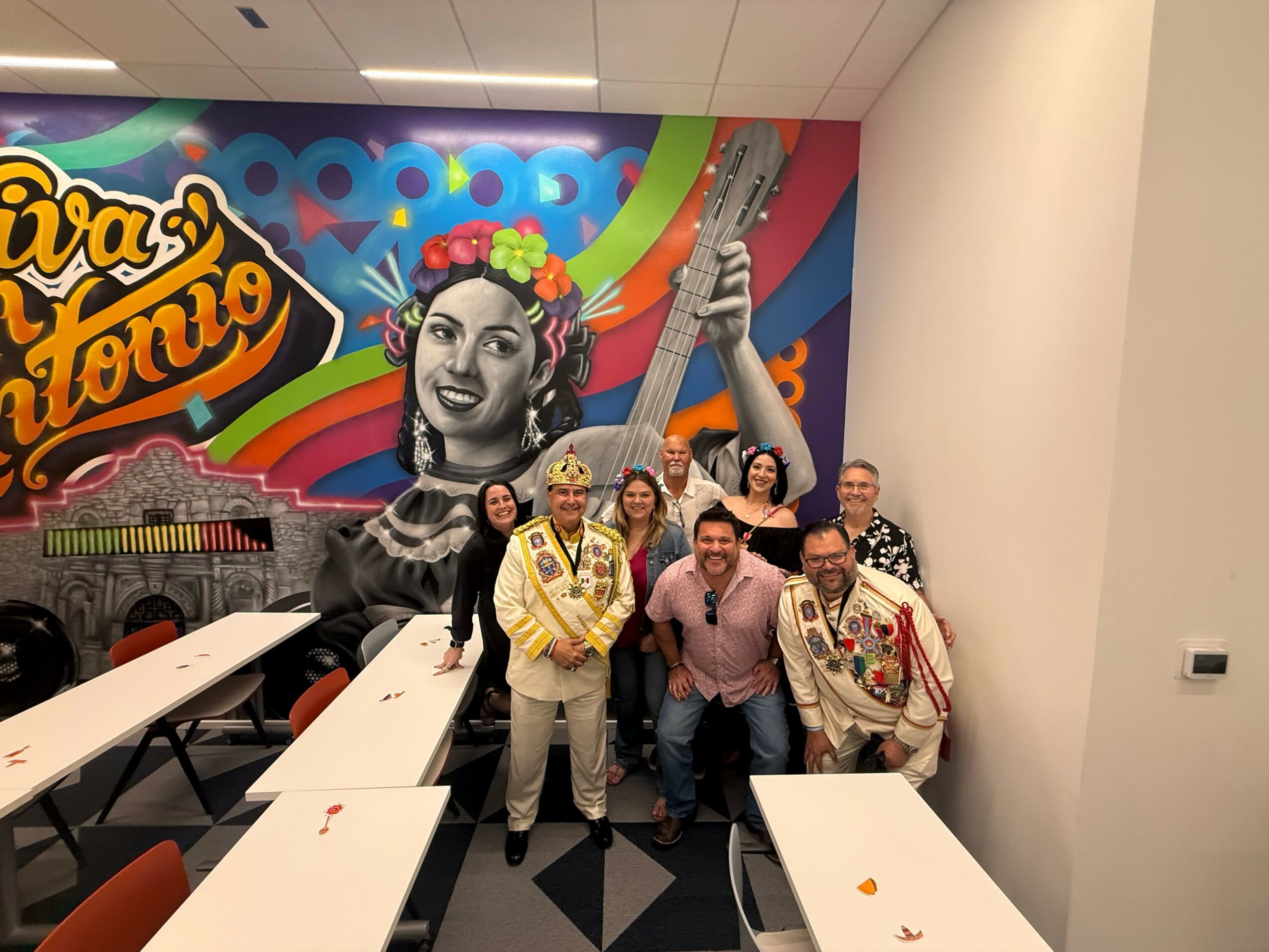 Group of seven people standing in front of a colorful mural with a woman playing a guitar, some smiling and posing.