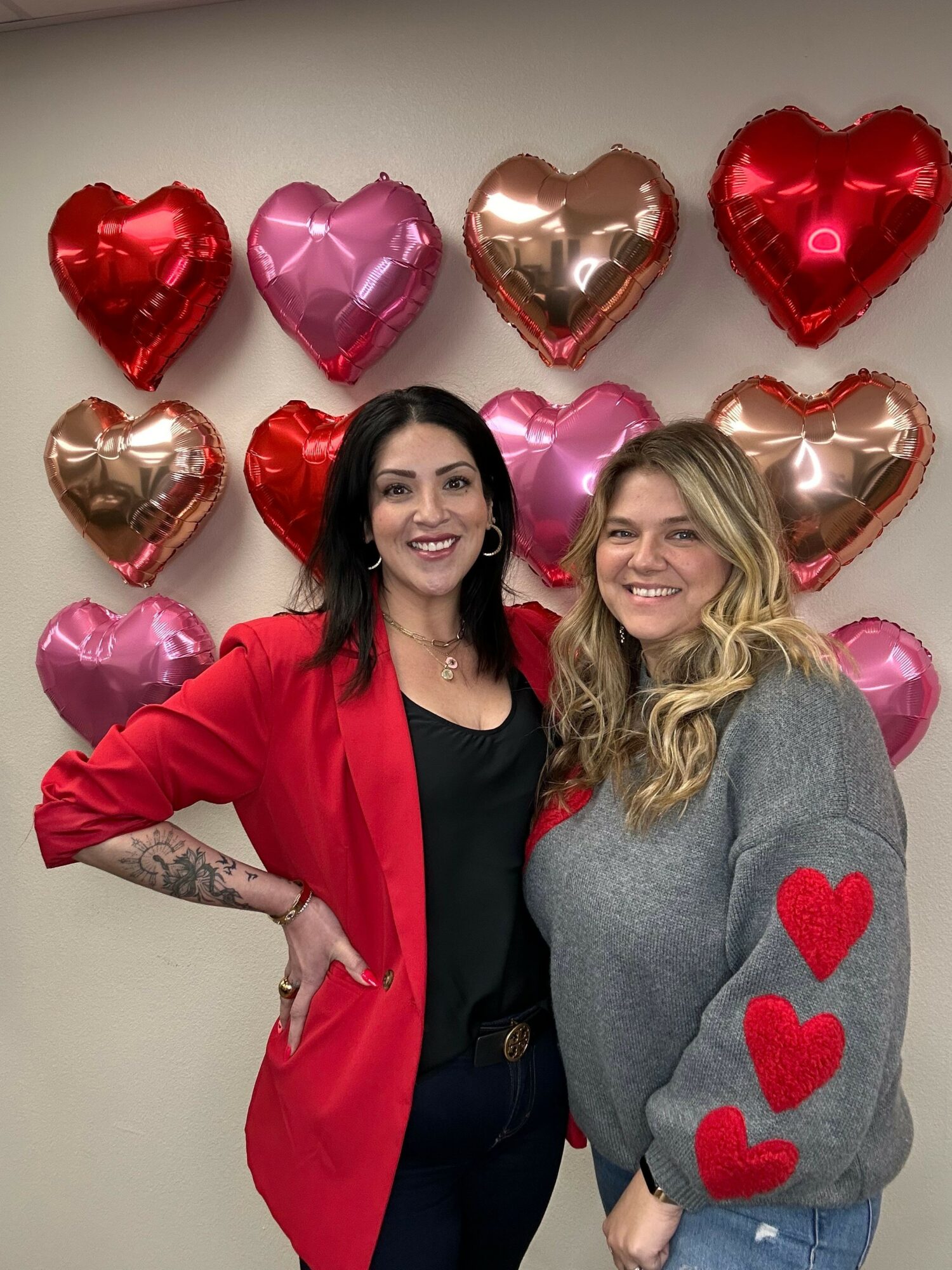 Two women smiling, surrounded by red, pink, and rose gold heart-shaped balloons, one wearing a gray sweater with red hearts, the other in a red jacket.