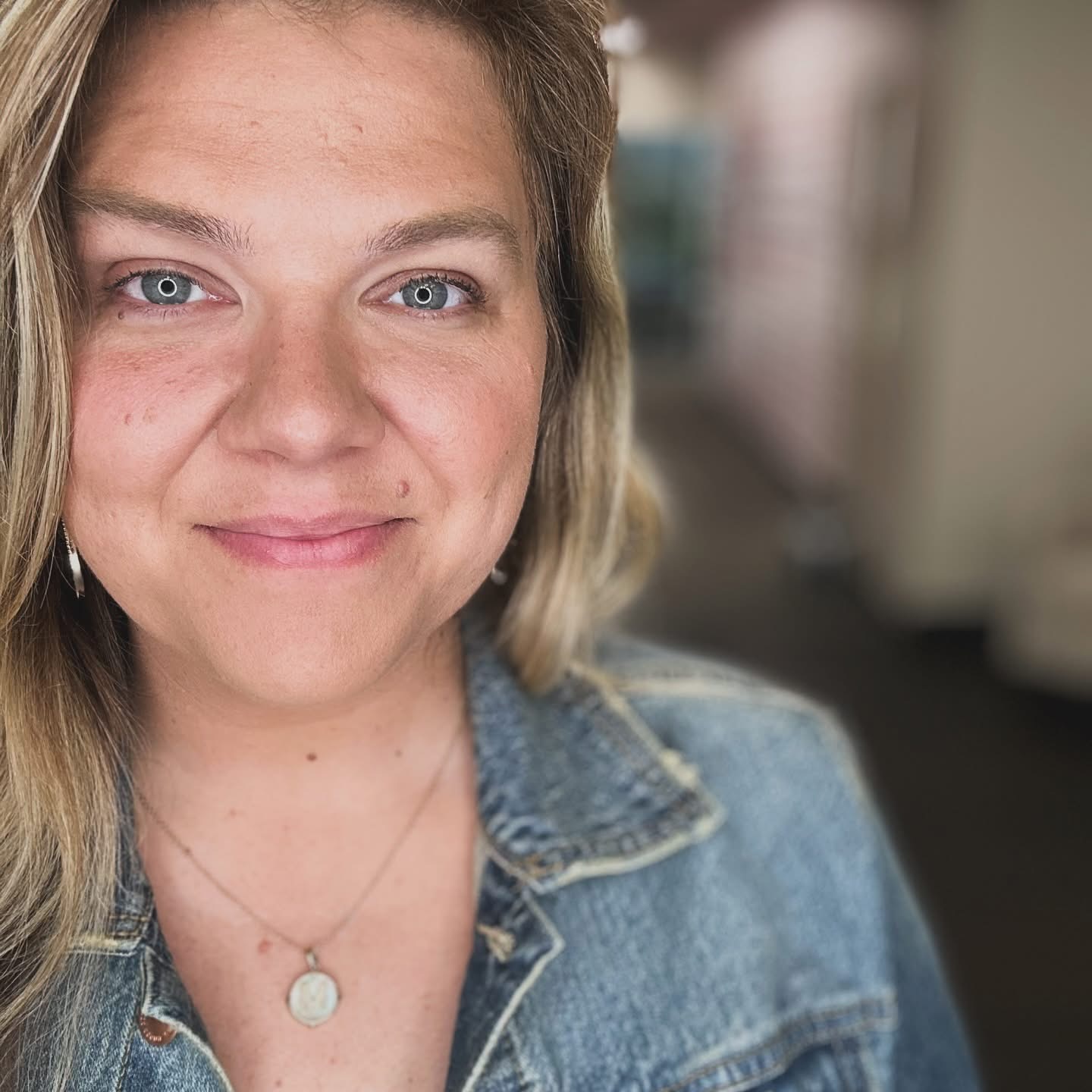 Close-up of a smiling woman with blonde hair and blue eyes, wearing a denim jacket and a necklace.