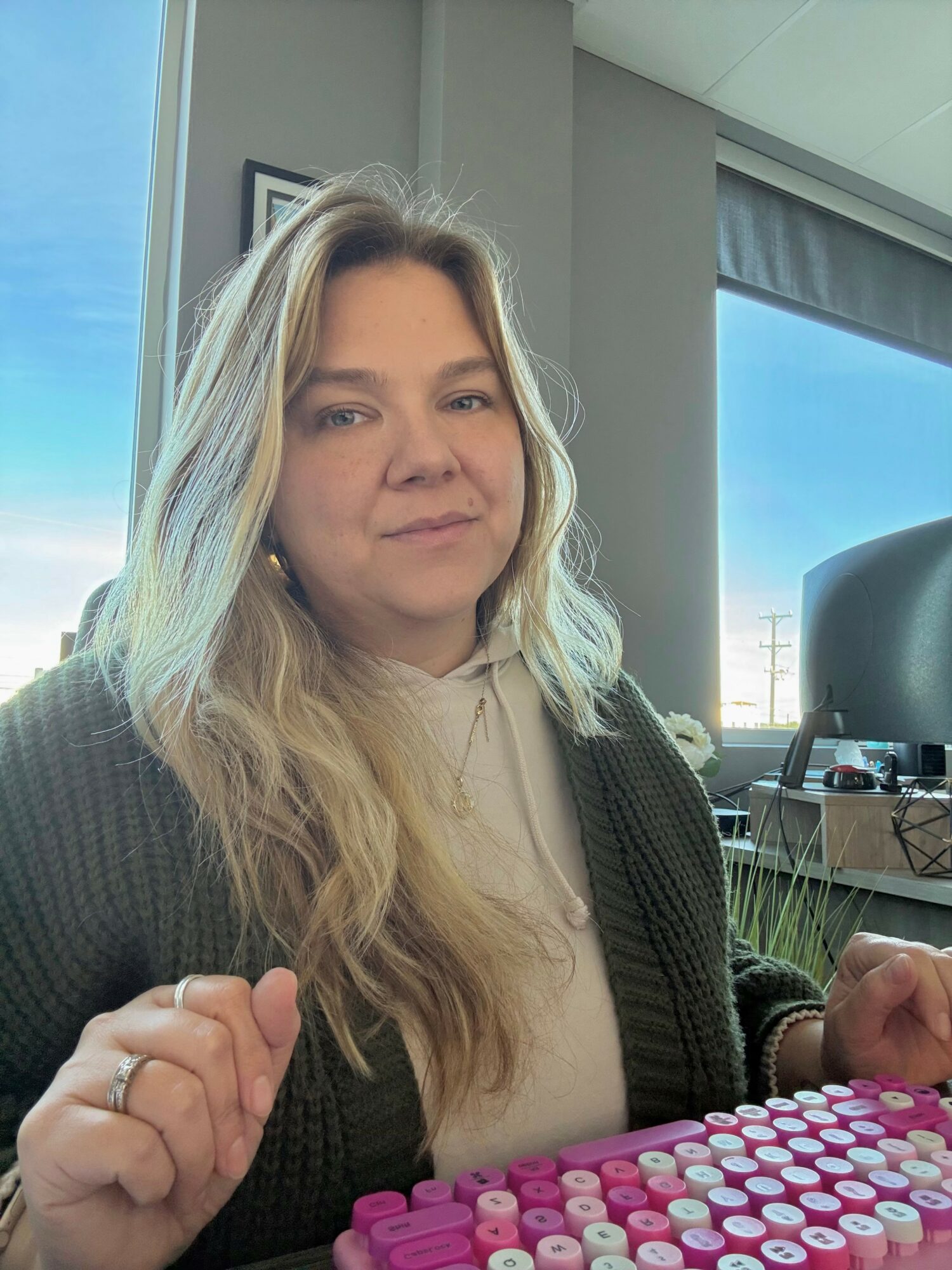 Young woman with long wavy blonde hair sitting indoors, wearing a beige top and dark cardigan, with a window and blue sky behind her.