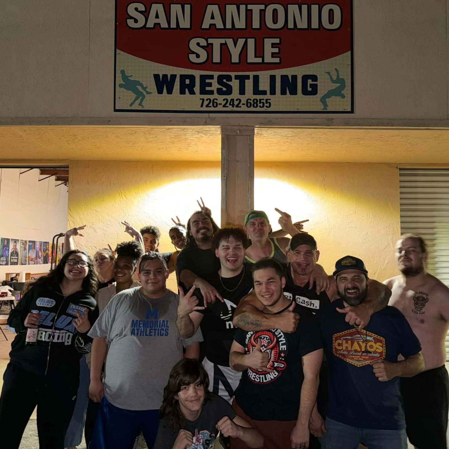 Group of people posing under a sign that reads 'San Antonio Style Wrestling' with a phone number, in an indoor setting.