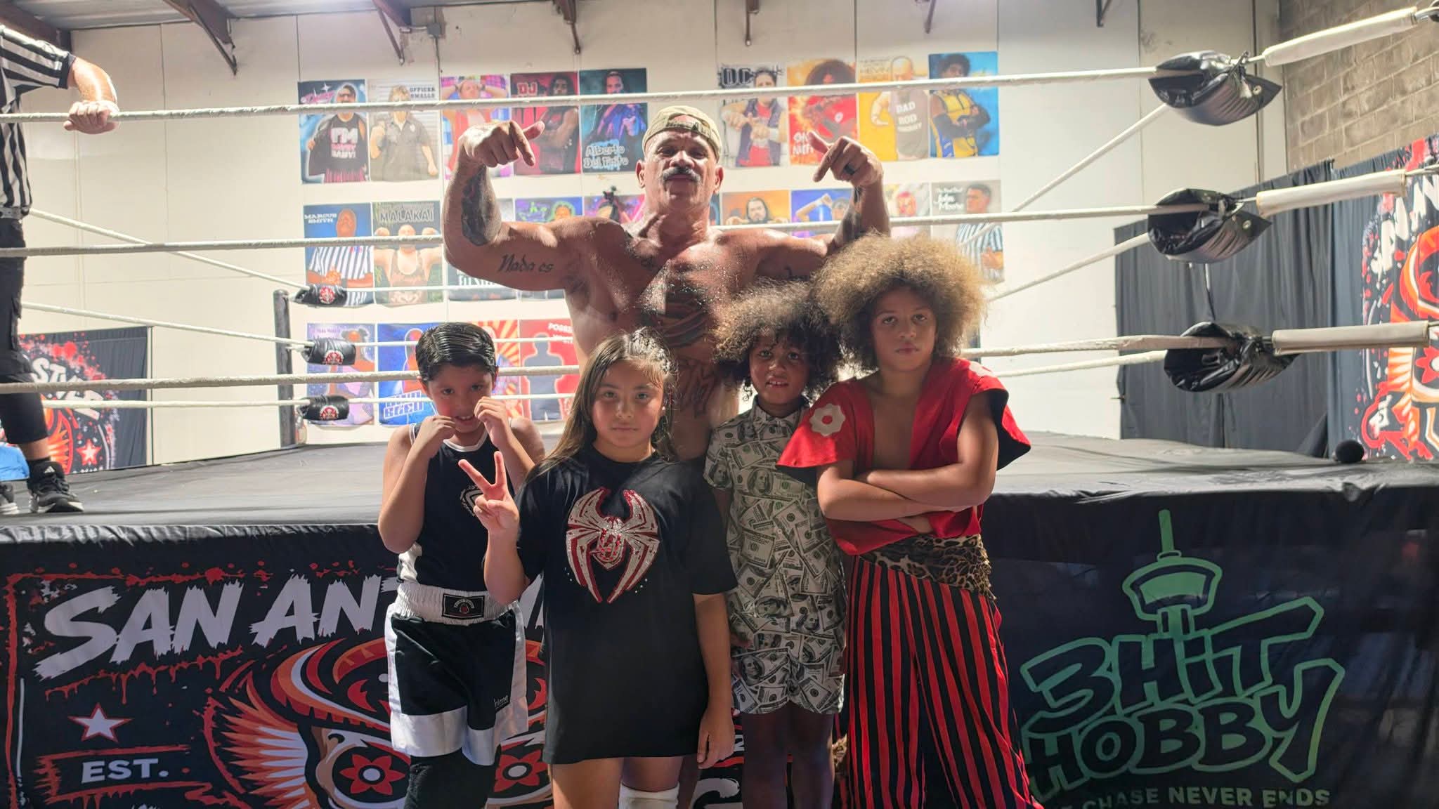 Group of five children and a man in a wrestling ring, with posters on the wall behind them.