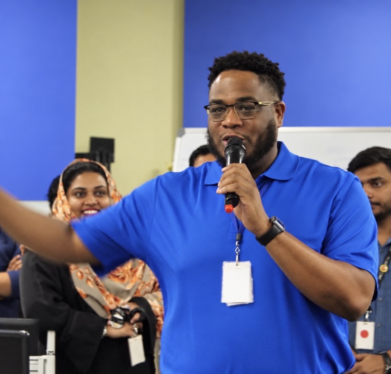 Man speaking into microphone during event, with smiling audience members in background, colorful walls behind him.