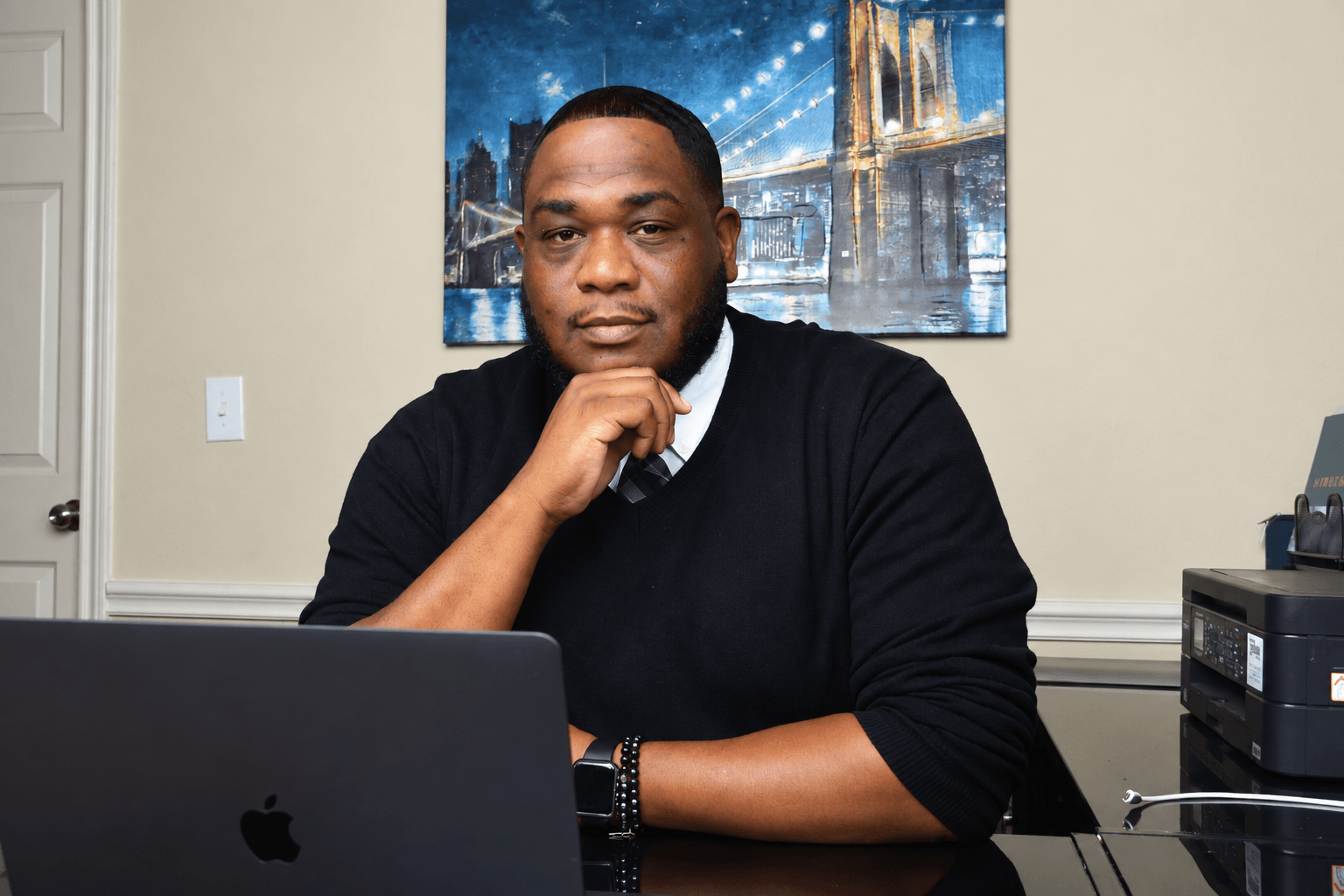 Man sitting at desk with laptop, hand on chin, in an office with cityscape artwork on wall.