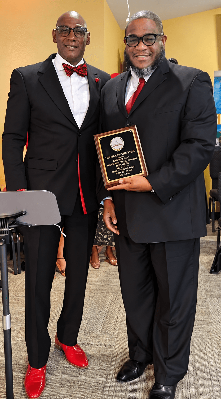 Two men in suits, one holding a plaque, standing indoors with a yellow wall background.