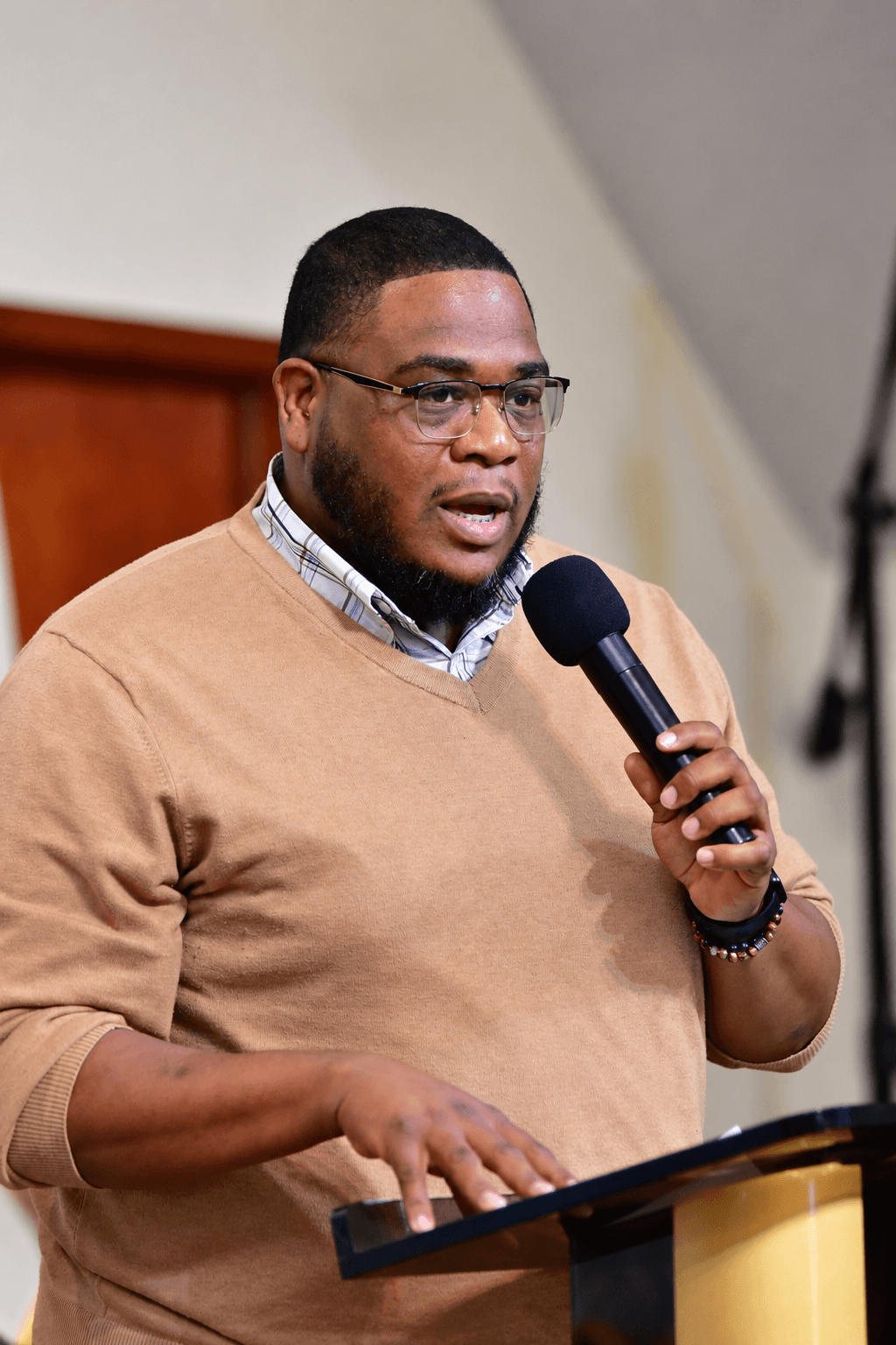Man speaking into a microphone at a podium, wearing glasses and a beige sweater, with a wooden background.