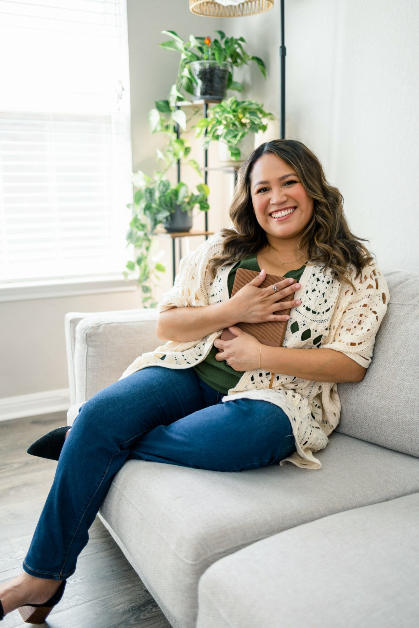 Woman sitting on sofa, smiling, holding a phone, in a bright room with plants and window blinds