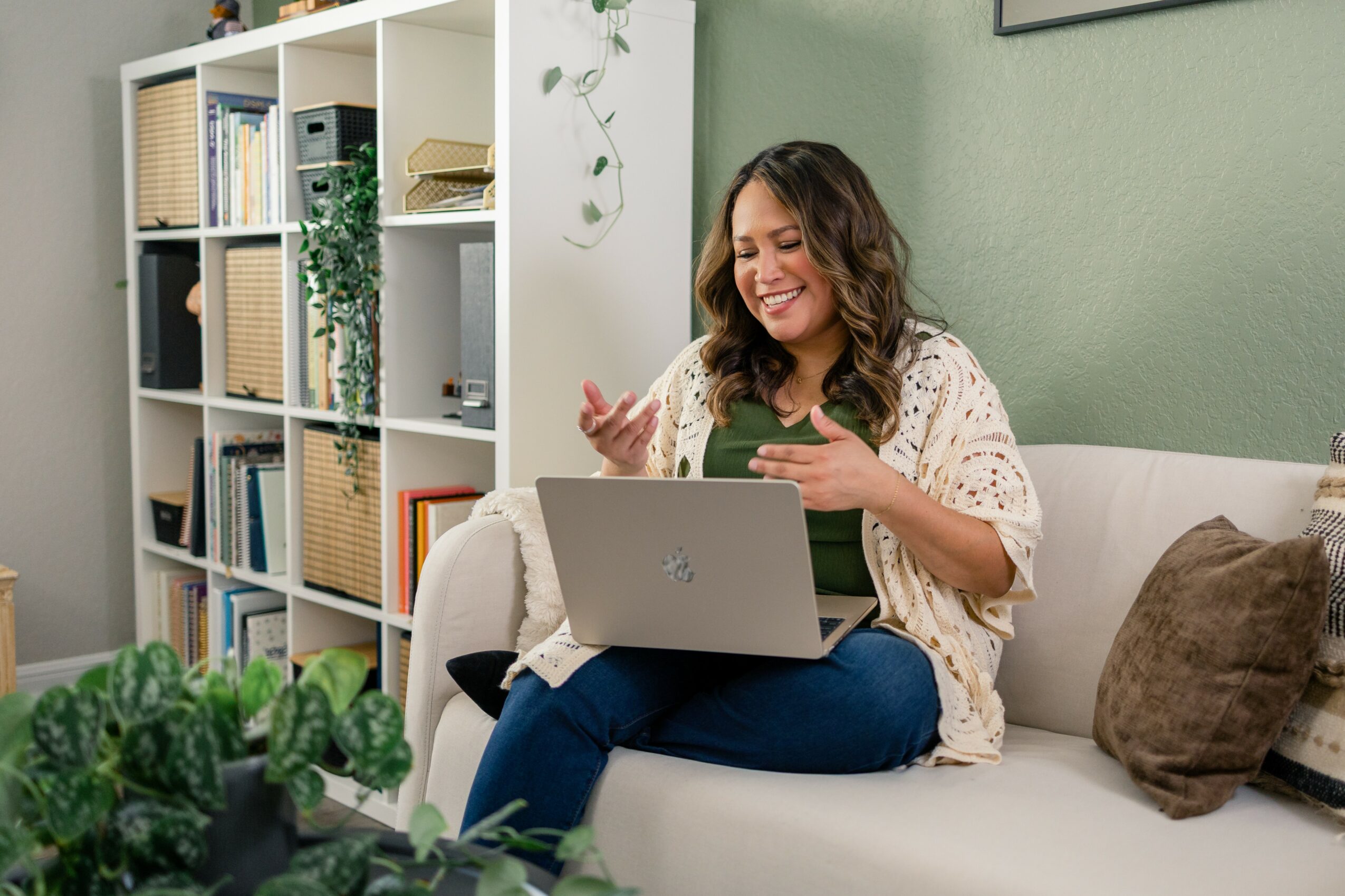 Woman sitting on a sofa using a laptop, smiling, in a room with a bookshelf and green wall.