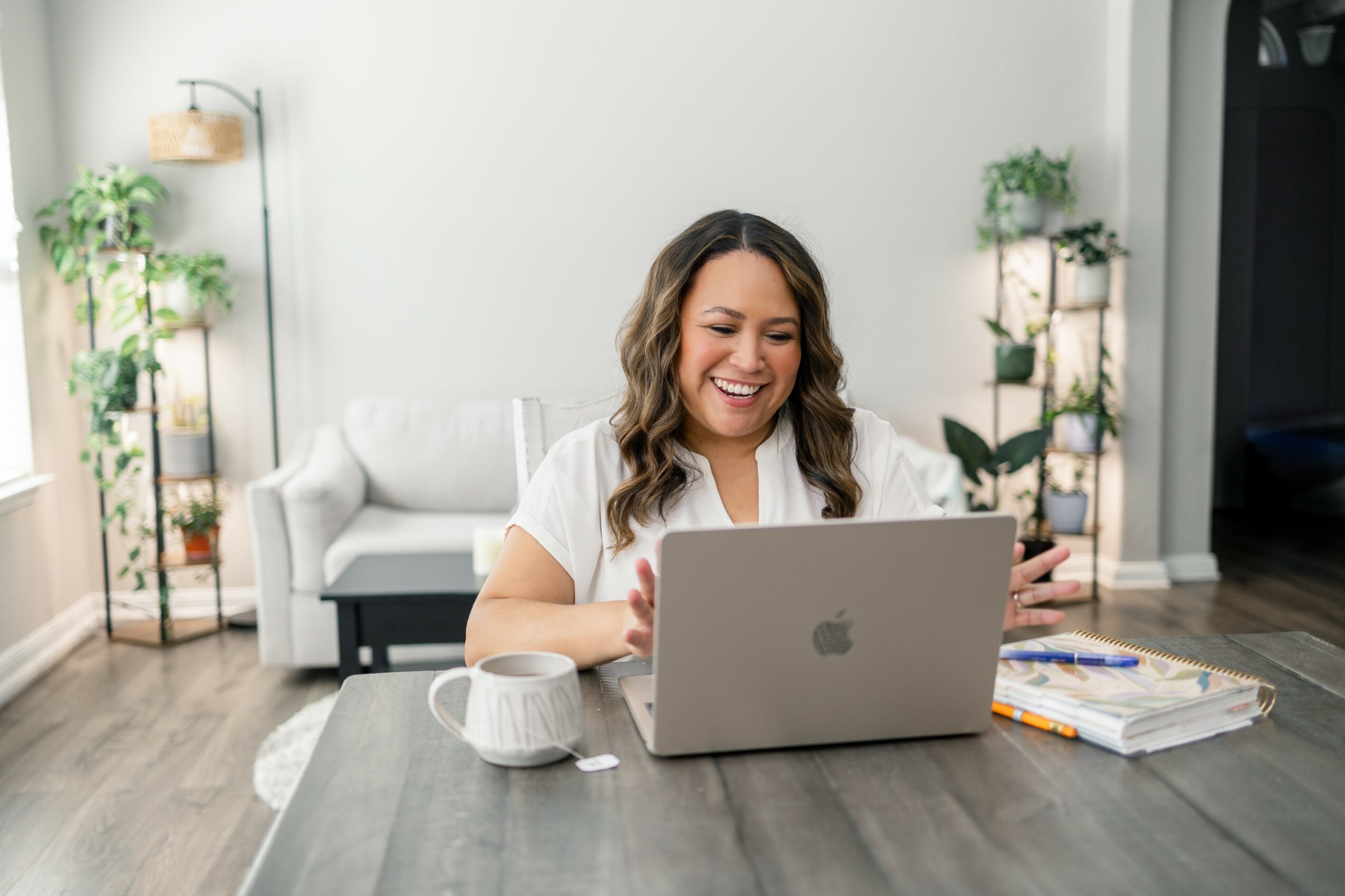 Woman smiling at laptop on table in bright living room with plants and sofa