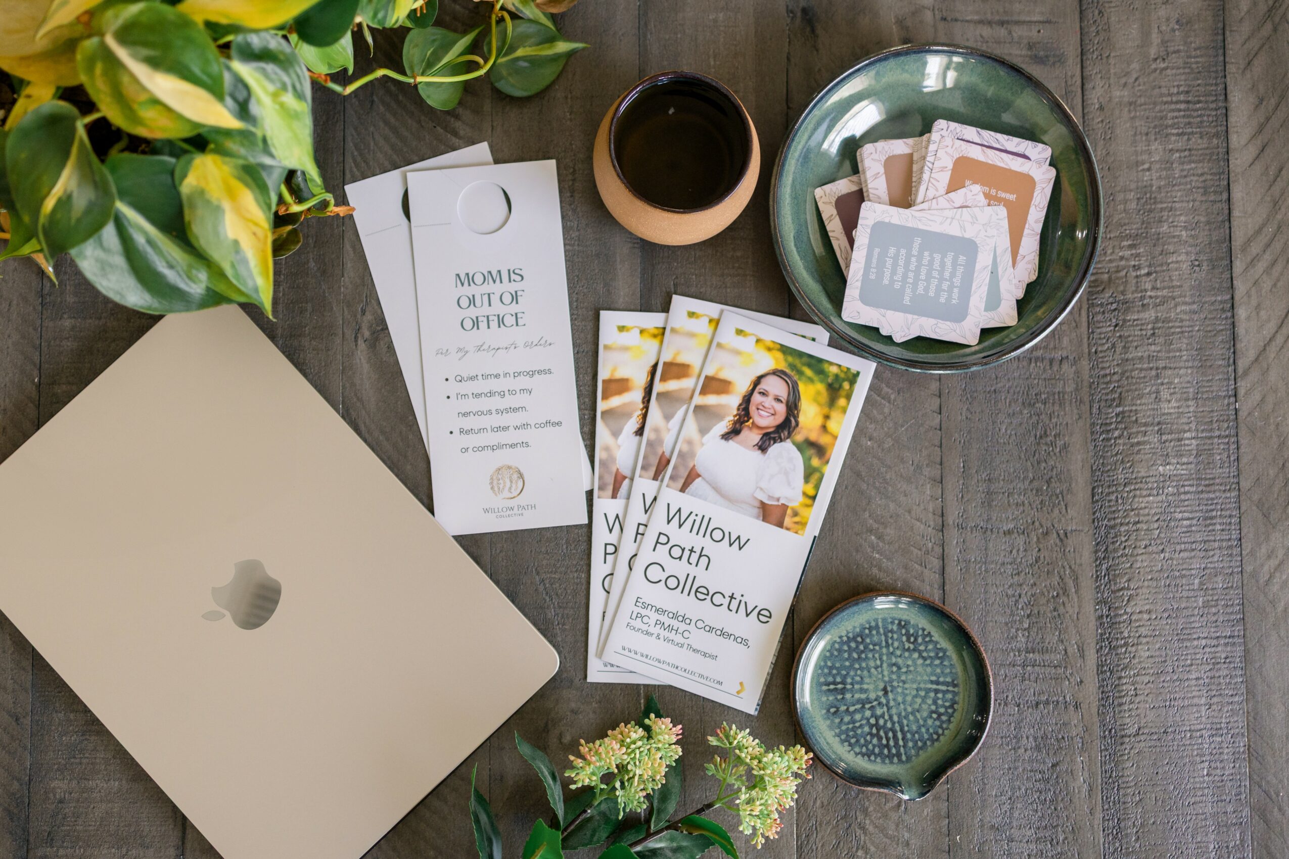 Open laptop on wooden table with plant, coffee, and printed materials nearby.