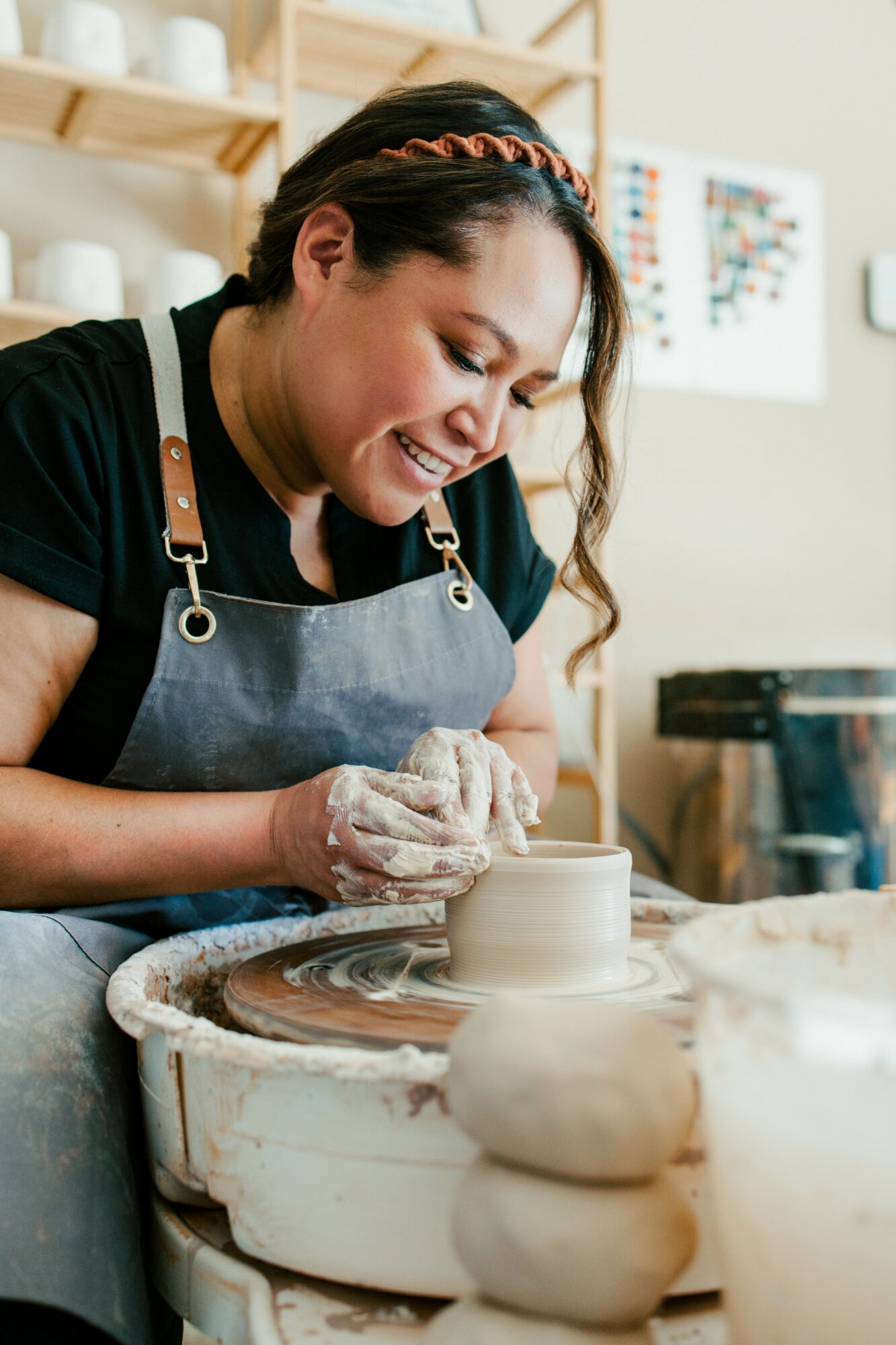 Woman shaping a ceramic piece on a pottery wheel in a studio, smiling and wearing an apron.