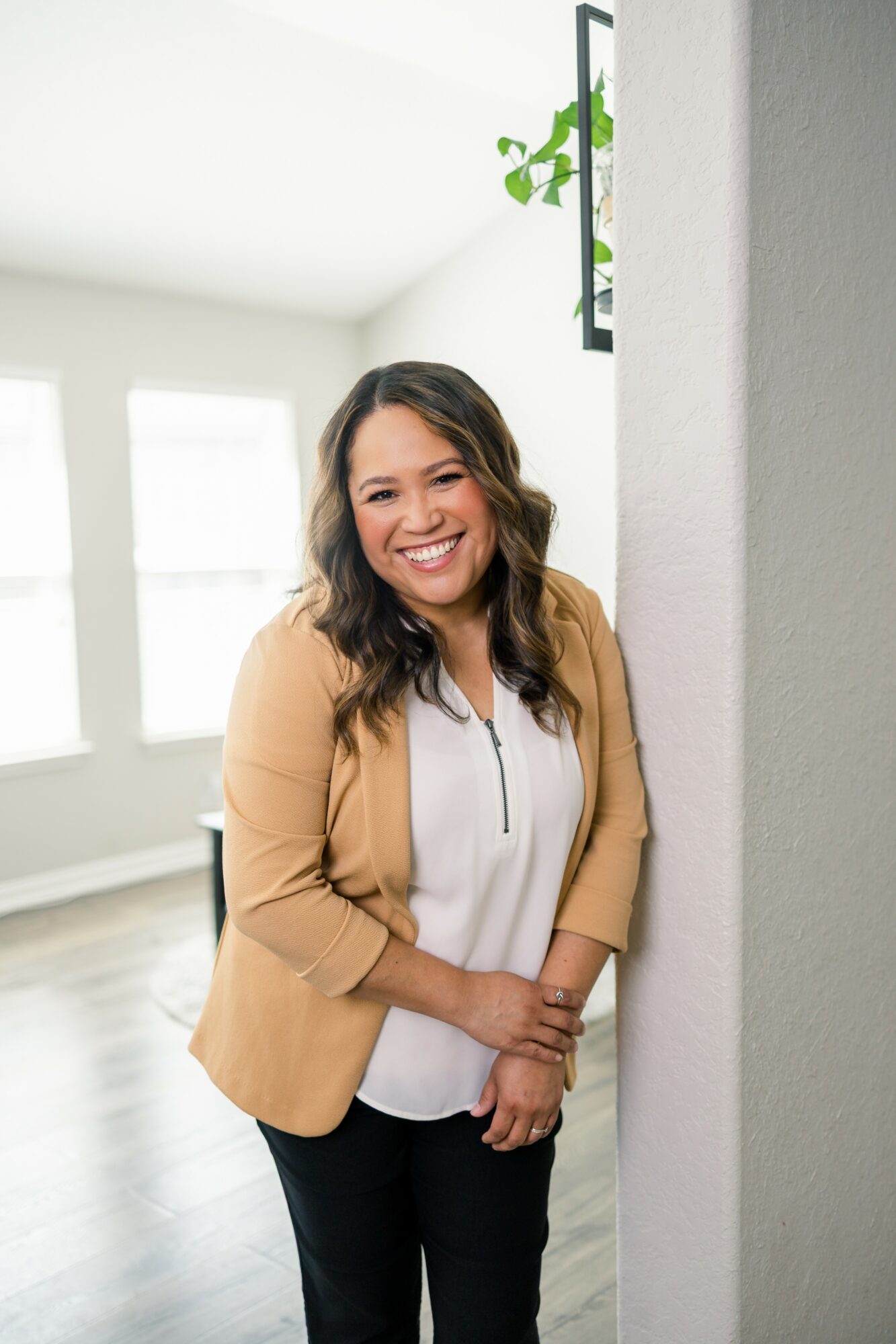 Woman smiling, standing indoors near a wall with windows in the background.