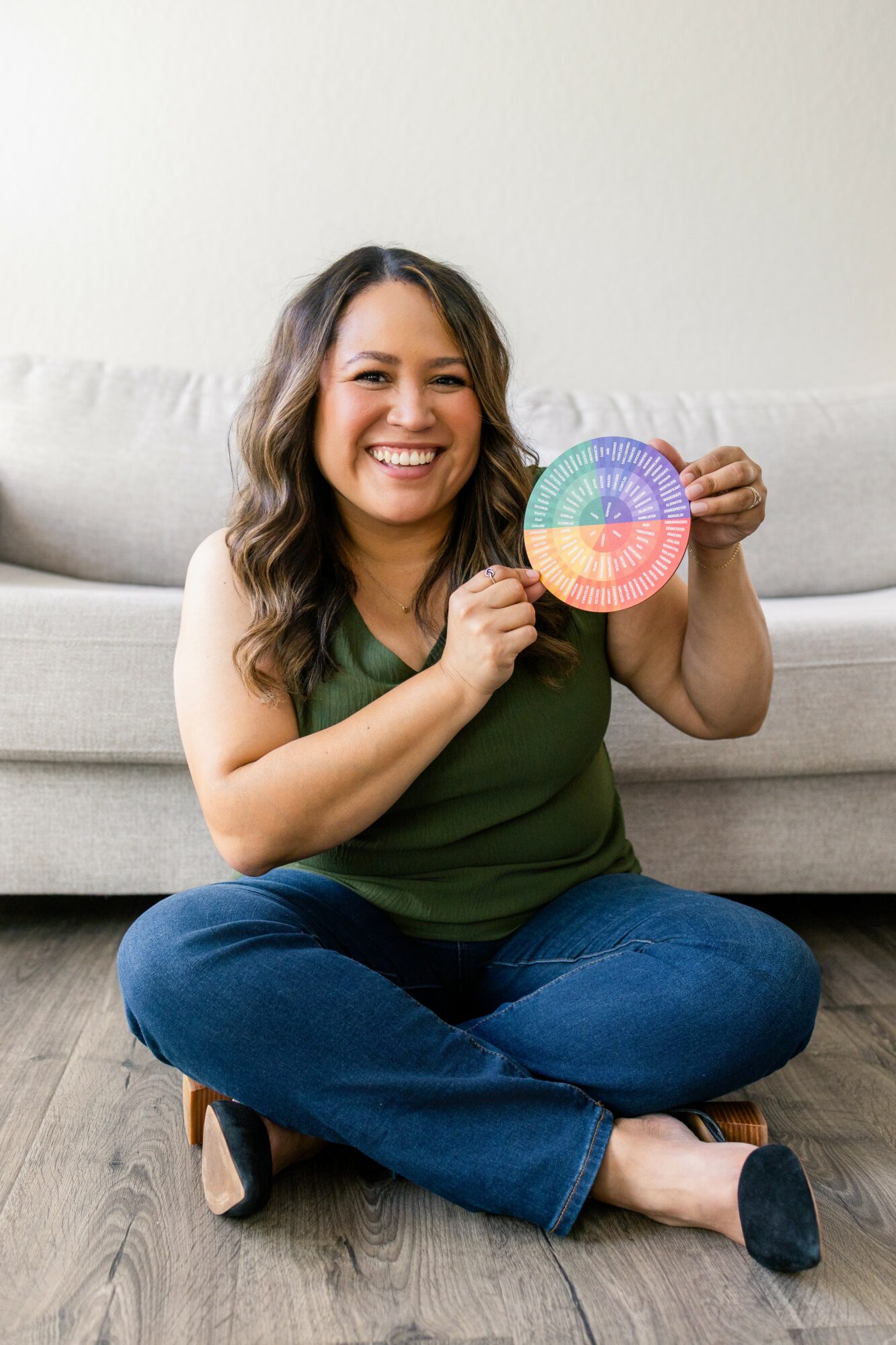 Woman sitting on floor holding a colorful circular object, smiling, in front of a light-colored sofa.