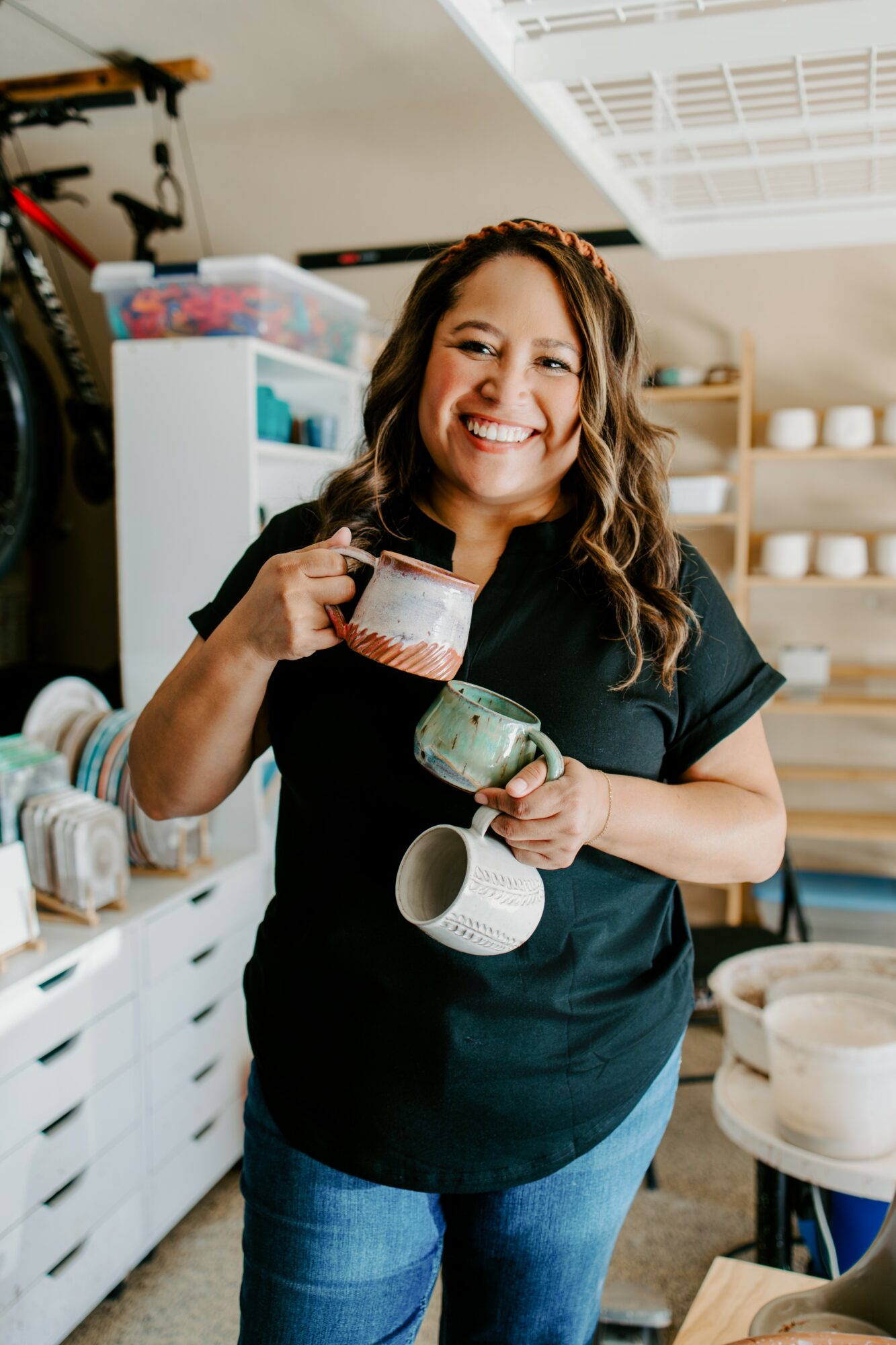 Woman smiling and holding a mug in a craft or pottery studio with shelves and supplies behind her.