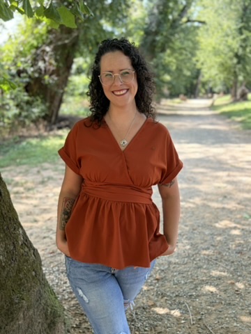 Woman with curly dark hair and glasses smiling outdoors in a park, wearing an orange top and blue jeans.