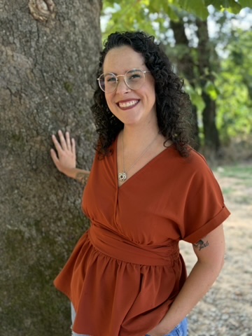 Woman with curly hair and glasses smiling outdoors, wearing a rust-colored dress, standing next to a tree.