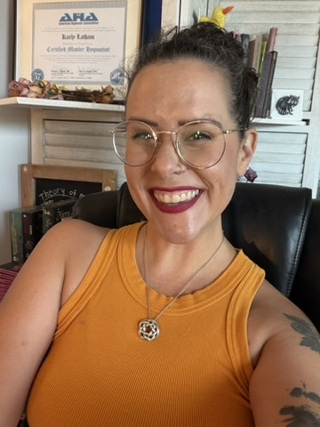 Smiling woman with glasses and a yellow top, sitting in a room with books and a certificate in the background.